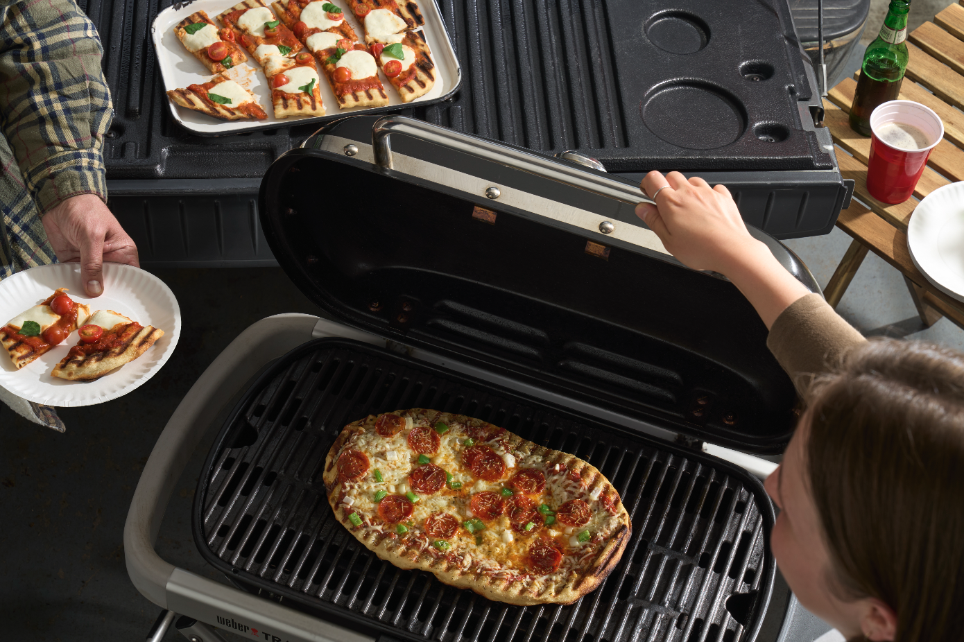 People grilling pepperoni and cheese pizza on an outdoor grill, with slices served on a paper plate.