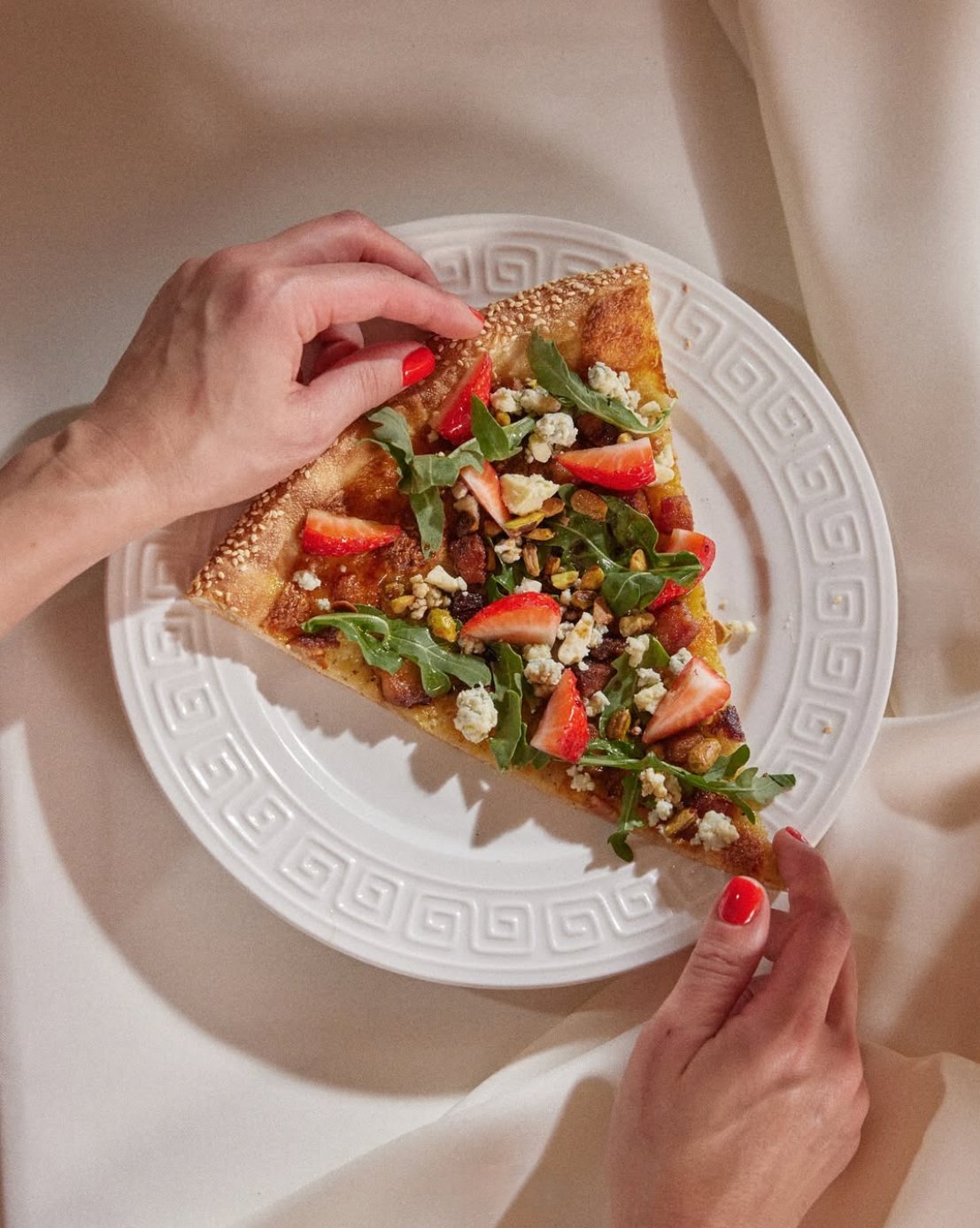 Person with red painted nails holding a slice of pizza topped with strawberries, cheese, arugula, nuts, and other ingredients on a decorative white plate.