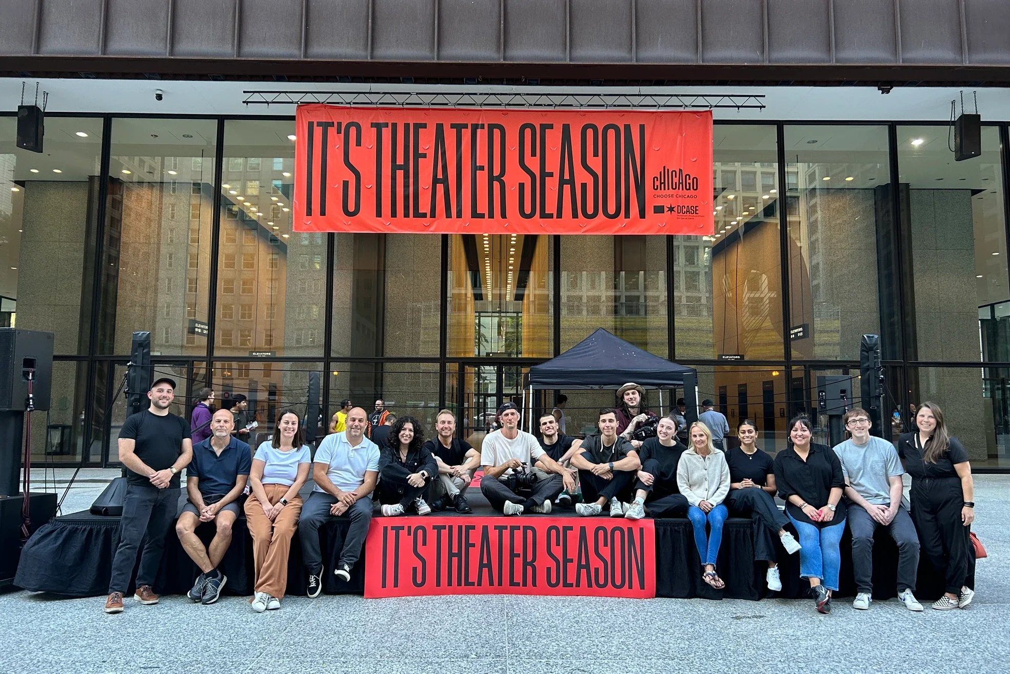 Group of people sitting and standing in front of a large building with glass windows, holding a red banner that reads 'IT'S THEATER SEASON' and a smaller banner with logos and the text 'chicago' and 'DCASE'.