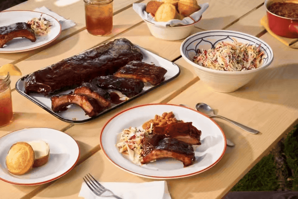 A barbecue meal including ribs, coleslaw, cornbread, and sides on a wooden table.