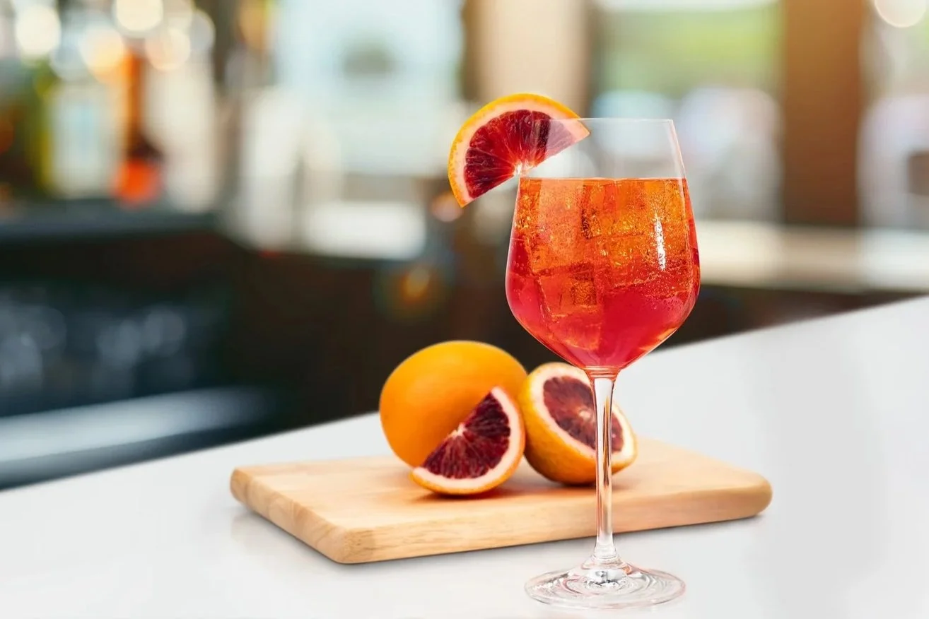 A glass of pink-colored cocktail with ice cubes and a slice of blood orange on the rim, placed on a white surface. There are whole blood oranges and blood orange wedges on a wooden cutting board in the background.