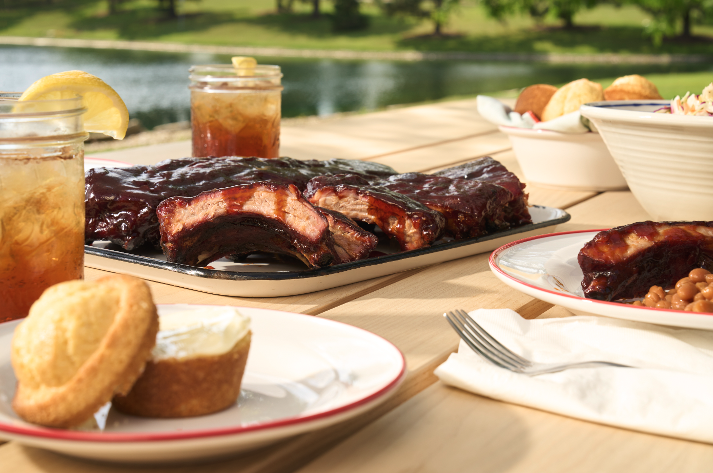 Barbecue ribs with baked beans, cupcakes, and lemonade on a picnic table outdoors near a lake.