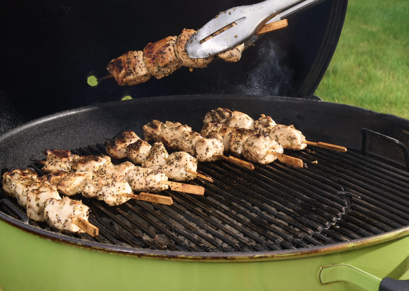 Chicken kebabs on a grill with one being flipped using tongs.