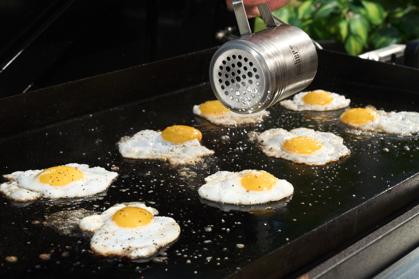 Fried eggs cooking on a griddle with a hand holding a metal seasoning shaker above them.