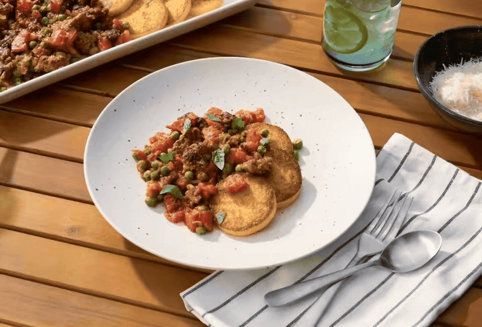 A white plate with fried green tomatoes topped with tomato and green onion relish, served on a wooden table next to a striped napkin with a fork and spoon, a glass of water with lime slices, and a bowl of salt or sugar, with a large dish of food partially visible in the background.