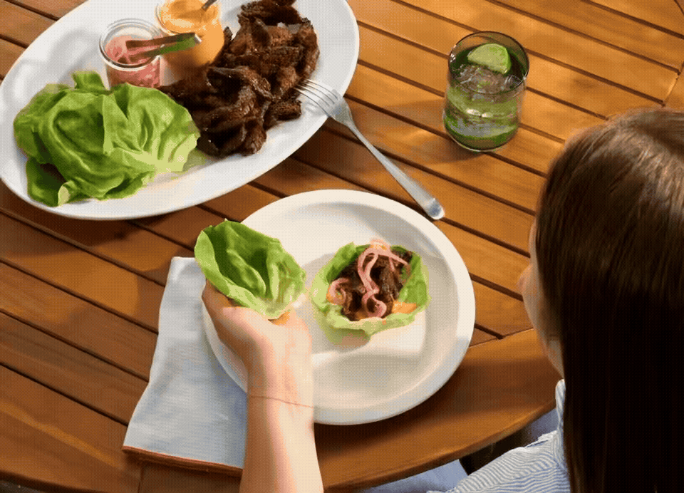 A person preparing a bowl of lettuce with sliced beef, onions, and sauce, with a plate of grilled beef, lettuce, and sauces, and a glass of lime and mint drink on a wooden table.