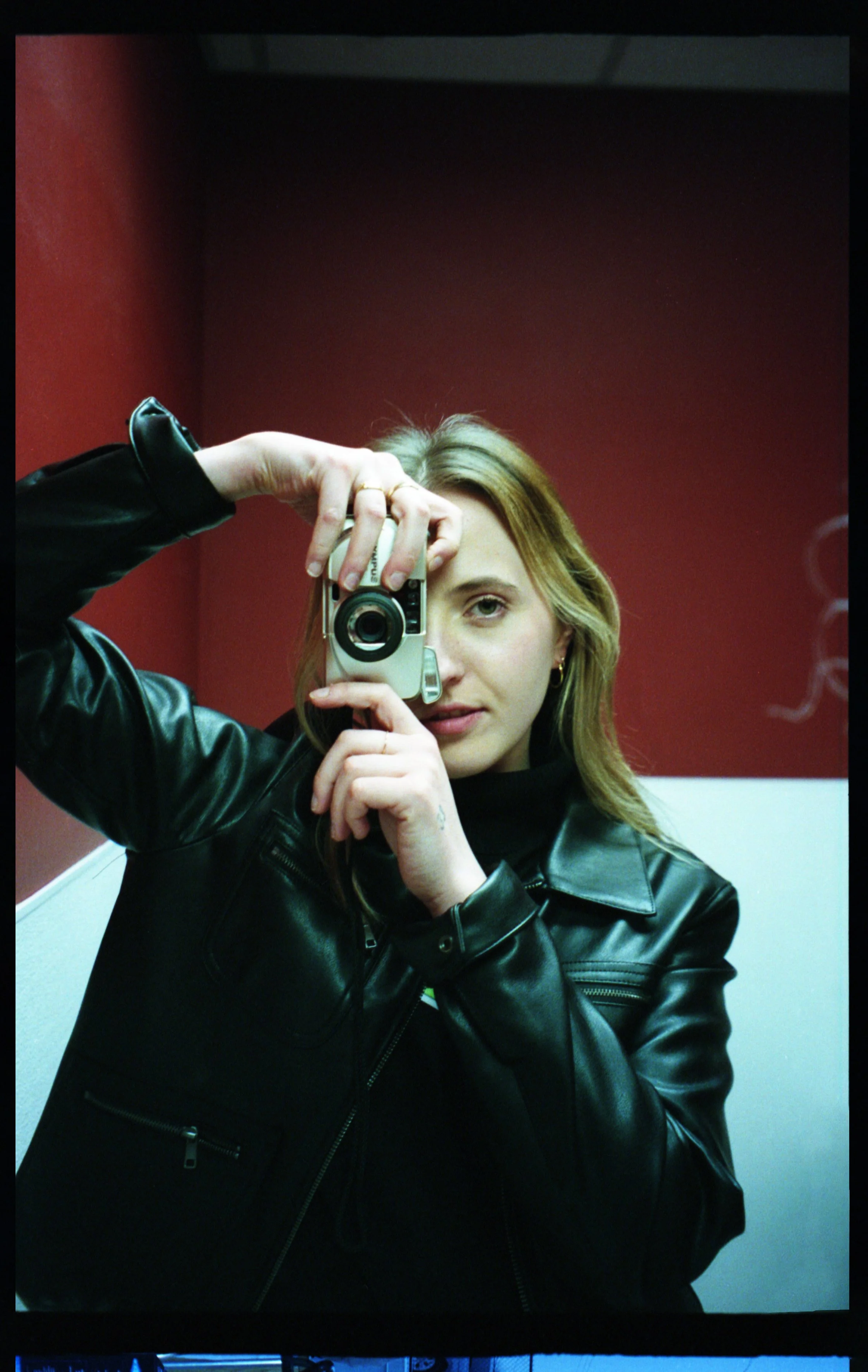 Woman taking a mirror selfie with a digital camera, wearing a black leather jacket, in a room with a red wall and white ceiling.