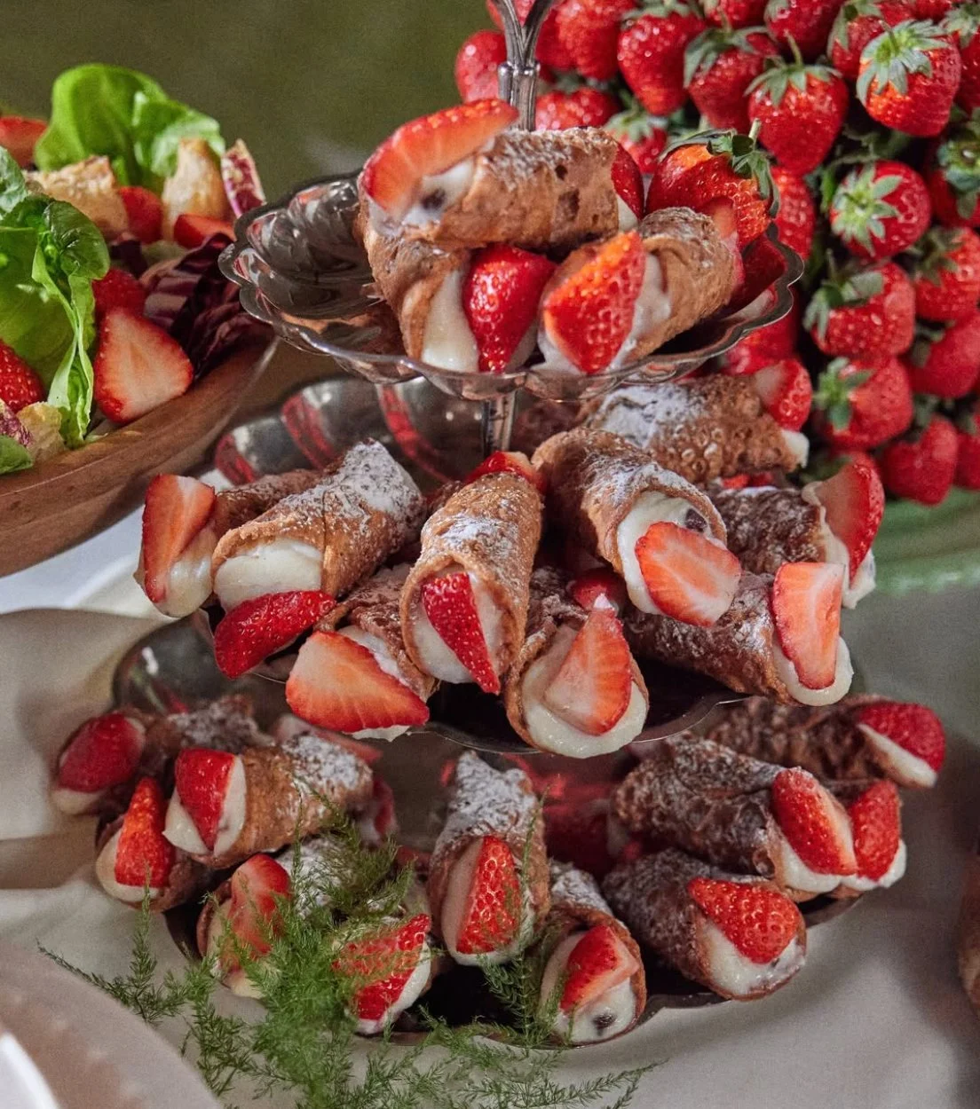 A three-tiered dessert stand holding cream-filled strawberries wrapped in waffle cones, topped with powdered sugar and fresh strawberries, with more strawberries and leafy greens as decoration.