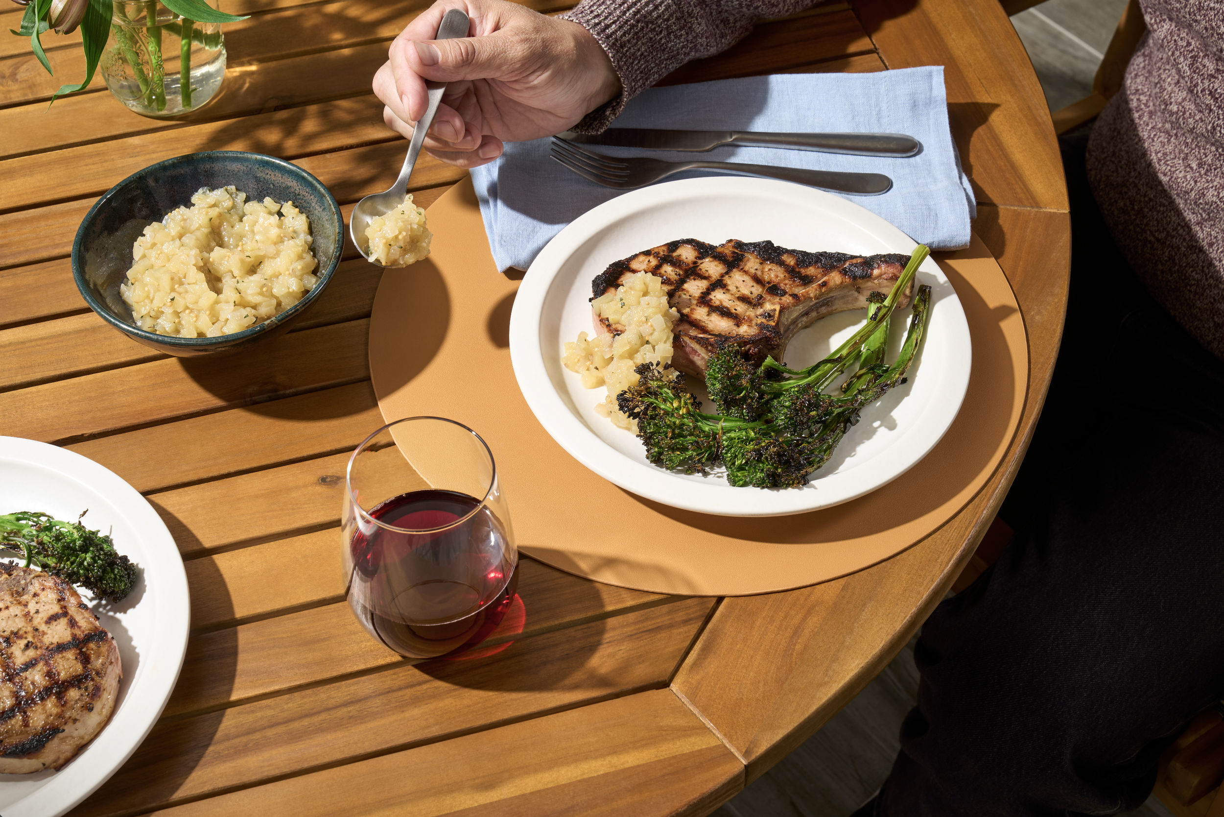 A person holding a spoon with potatoes, seated at a wooden table with a grilled steak, roasted vegetables, a glass of red wine, and a plant in a glass vase.