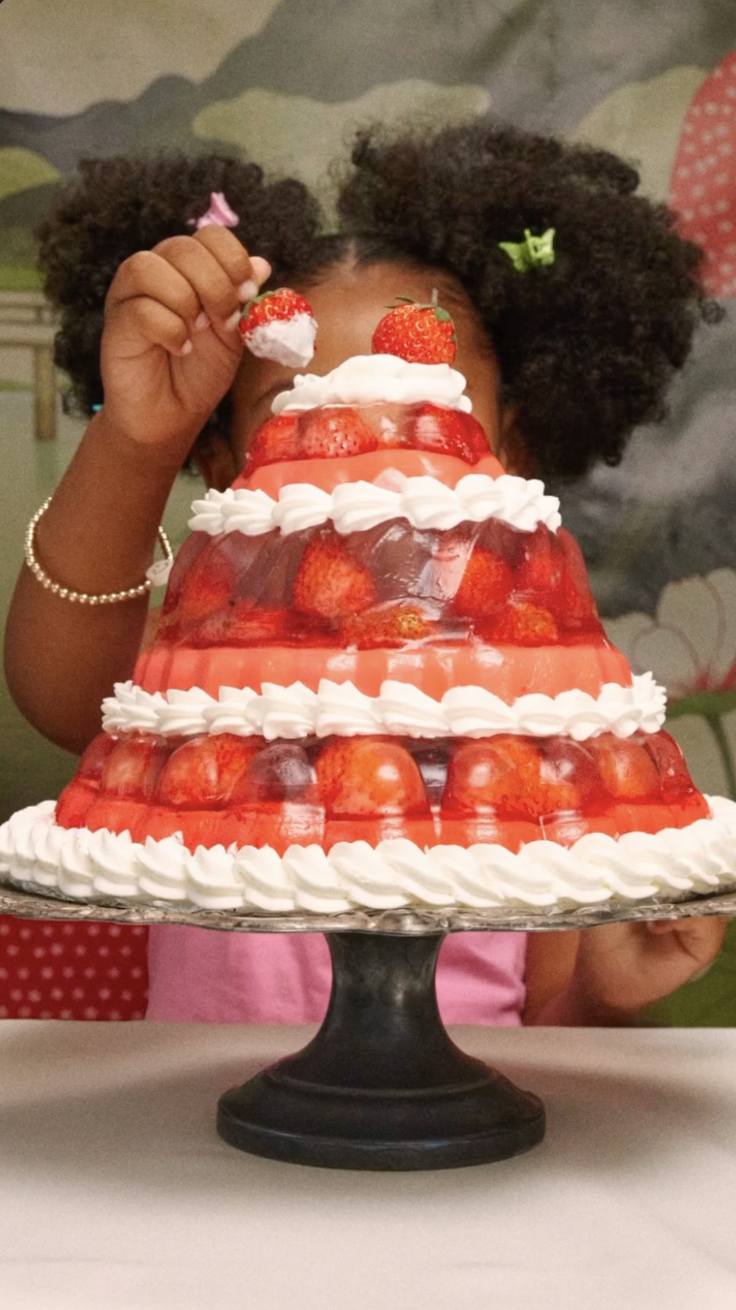 Child with curly hair and colorful hair accessories presents a multi-tiered strawberry-themed cake topped with strawberries, whipped cream, and pink icing, on a black cake stand.