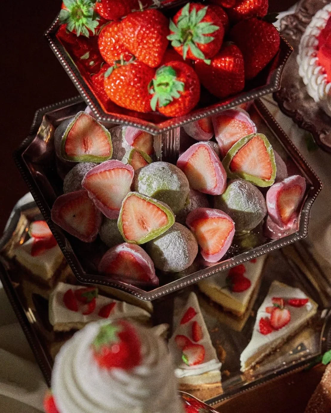 A three-tiered dessert stand with strawberries and chocolate-covered strawberries on the top, frozen mochi strawberries in the middle, and slices of cake with strawberries on the bottom.