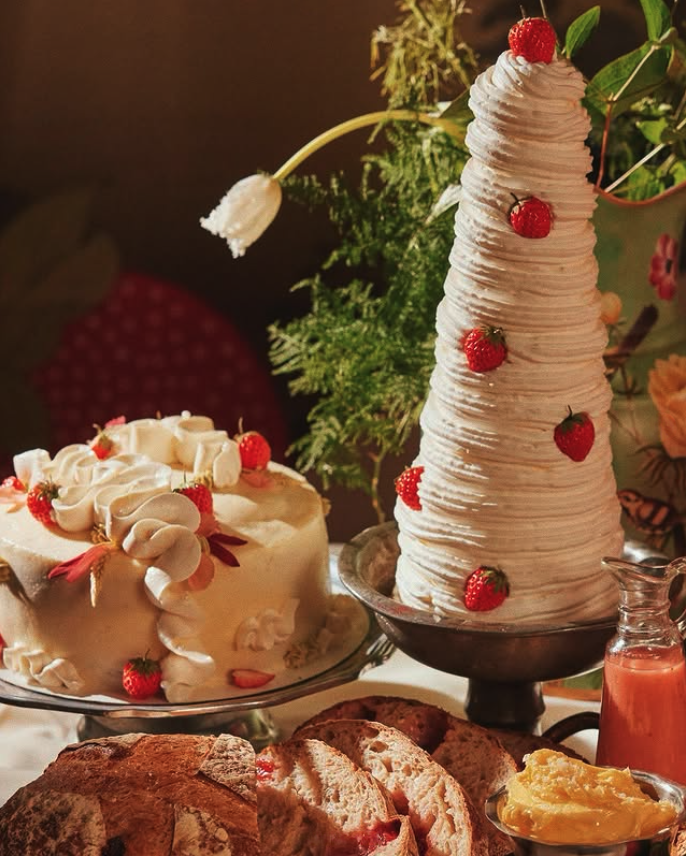 Dessert table with white frosted cake decorated with strawberries, a tall conical whipped cream dessert topped with a raspberry, uncut bread and a dish of butter, with a green plant and floral wallpaper in the background.