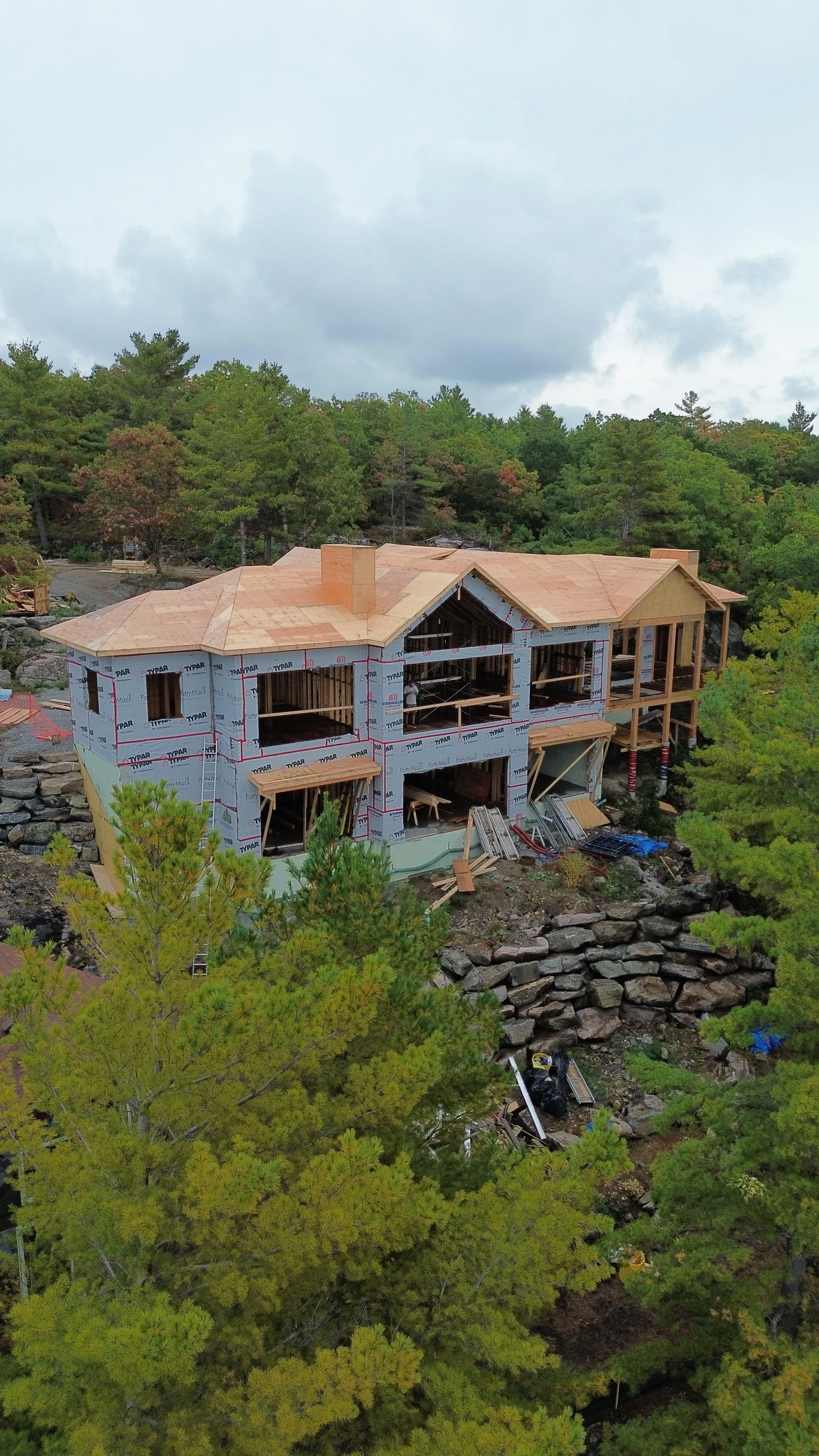 A house under construction on a hillside surrounded by trees with a cloudy sky overhead.