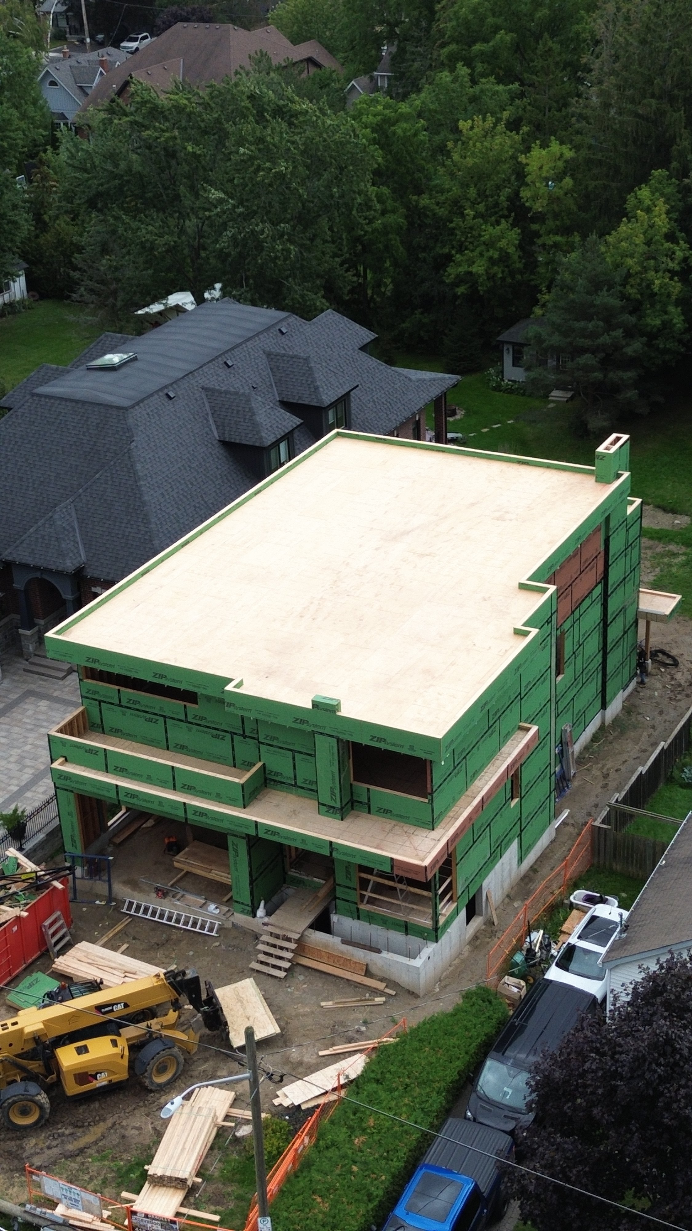 A house under construction with green exterior sheathing and no windows installed, surrounded by trees and grass.