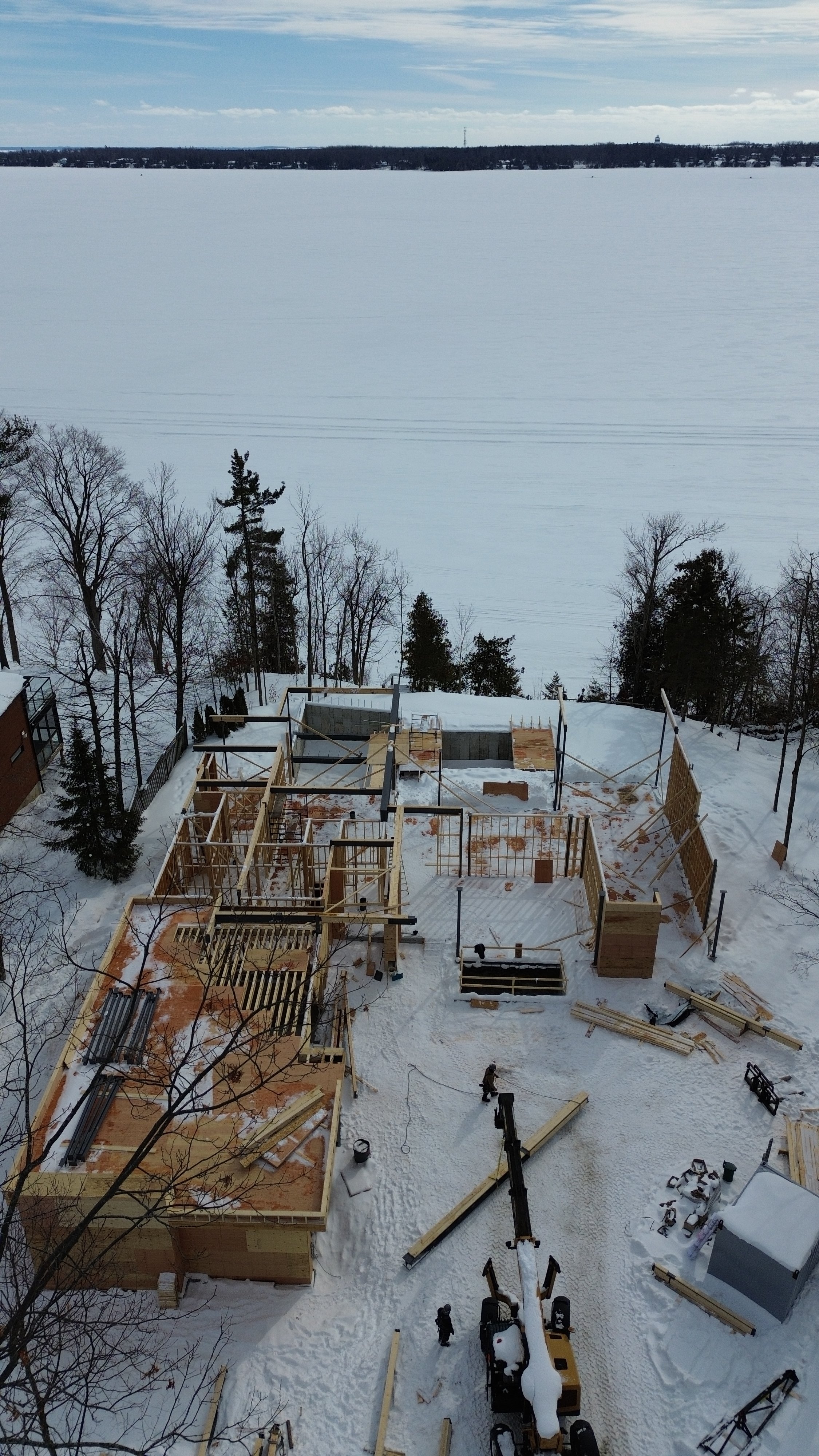 Construction site in a snowy landscape with a partially built wooden structure and a crane. Trees and a frozen lake are visible in the background.