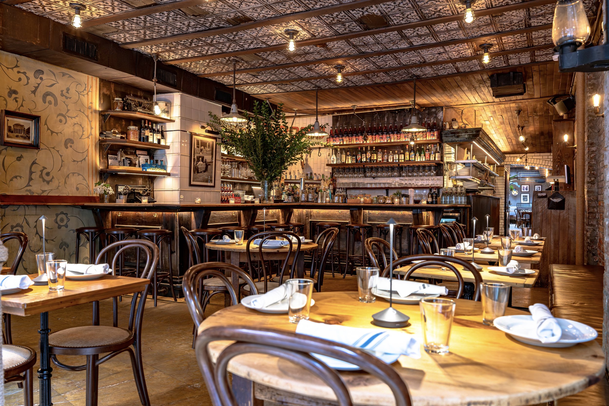Interior of a cozy restaurant with wooden tables and chairs, a bar area with bottles and glasses, warm lighting, and decorative ceiling tiles.