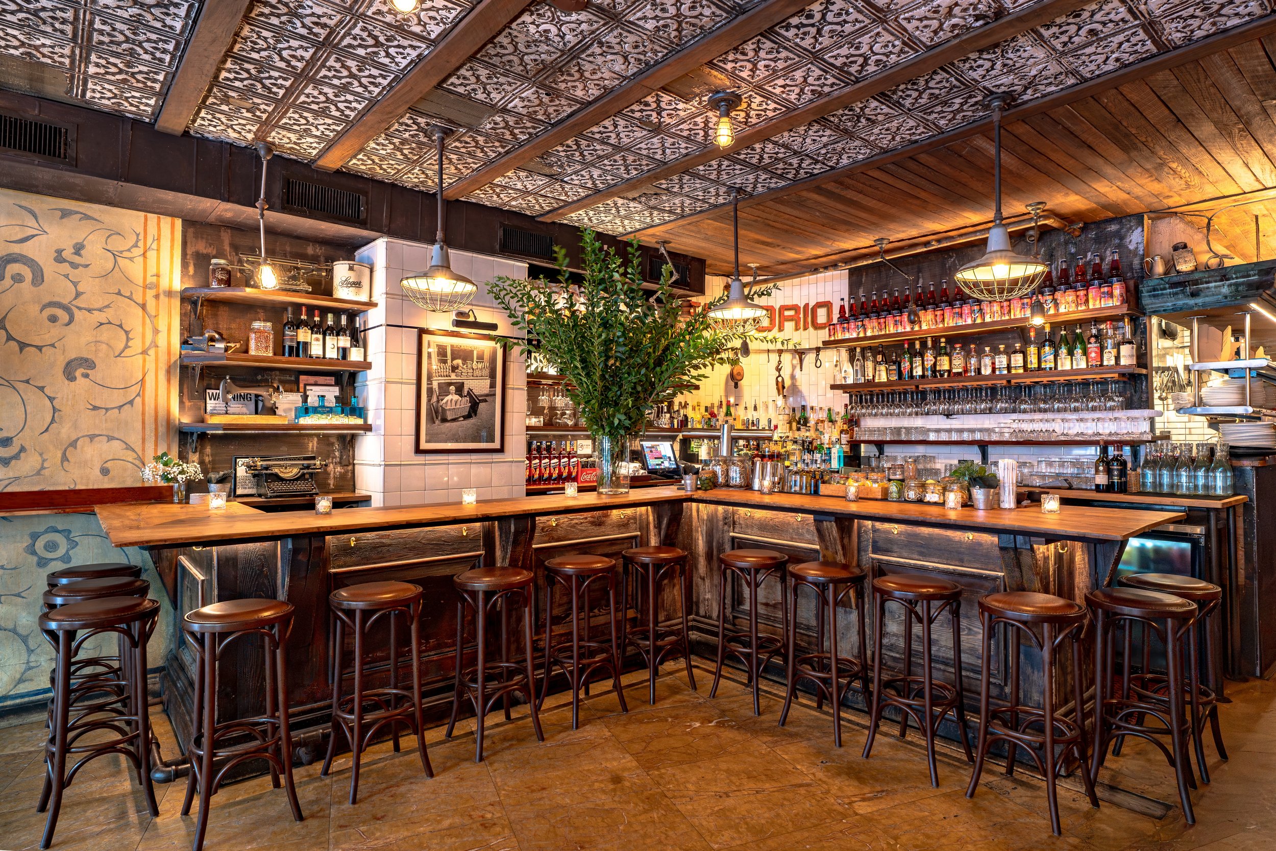 A cozy bar with a wooden counter, bar stools, shelves of liquor bottles, and decorative ceiling tiles in a rustic style.
