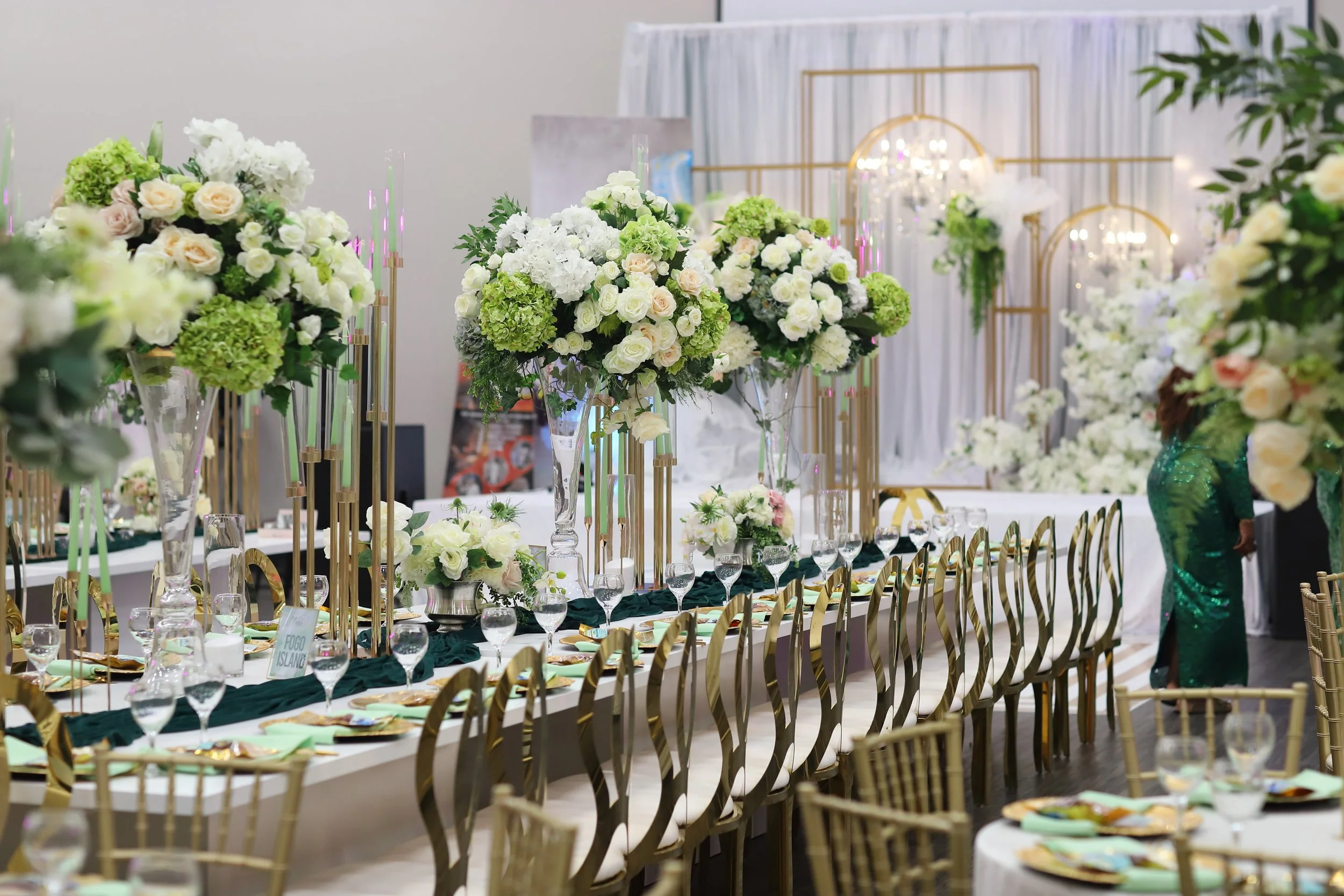 Elegant banquet table set for an event with tall floral centerpieces of white and green flowers, gold and black chairs, and decorative gold accents in the background.
