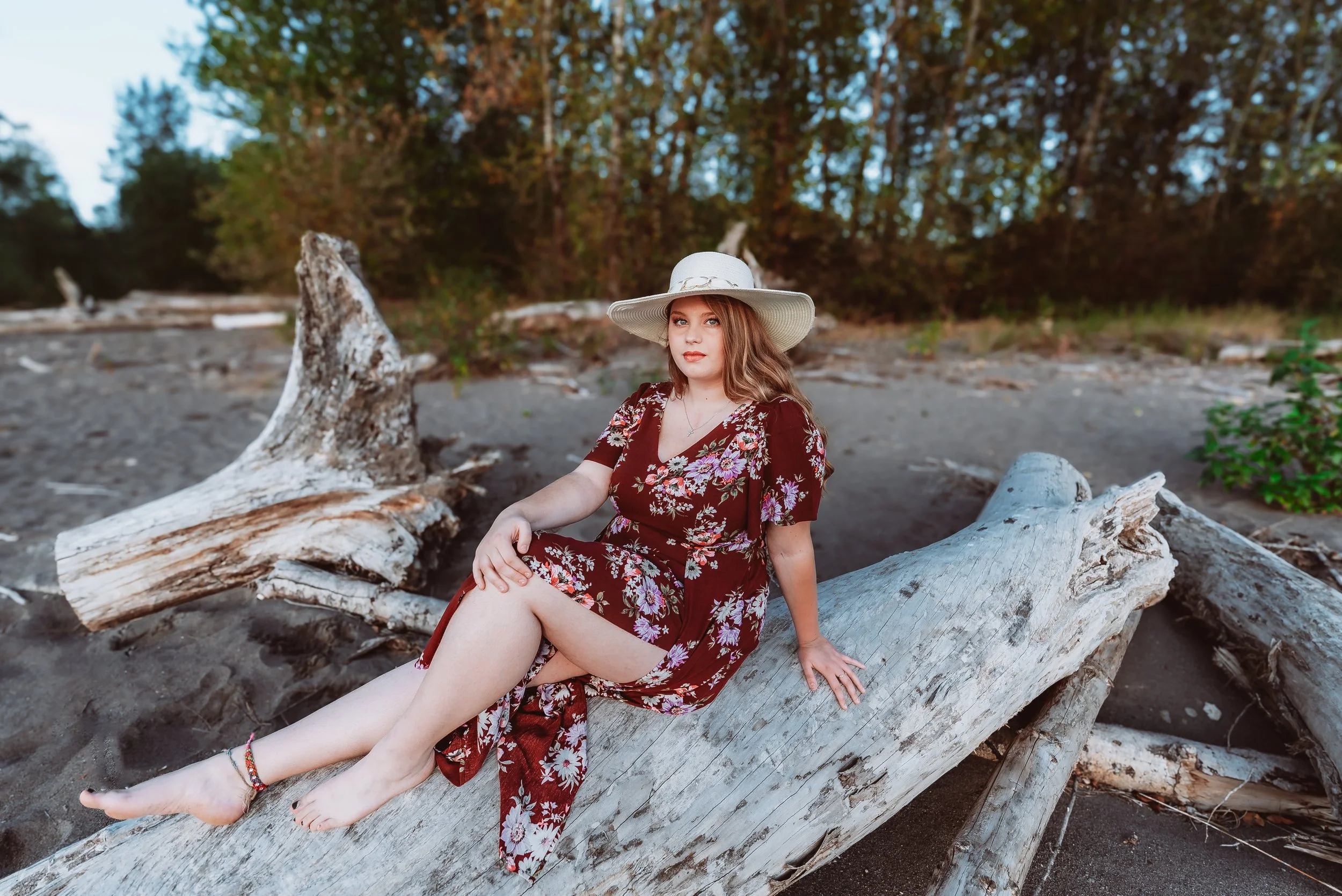 Woman in a floral dress and wide-brimmed hat sitting on a large Vancouver Washington senior photography session highlighting a graduating senior enjoying a calm, candid moment during an outdoor beach portrait shoot. log on a sandy beach with trees in