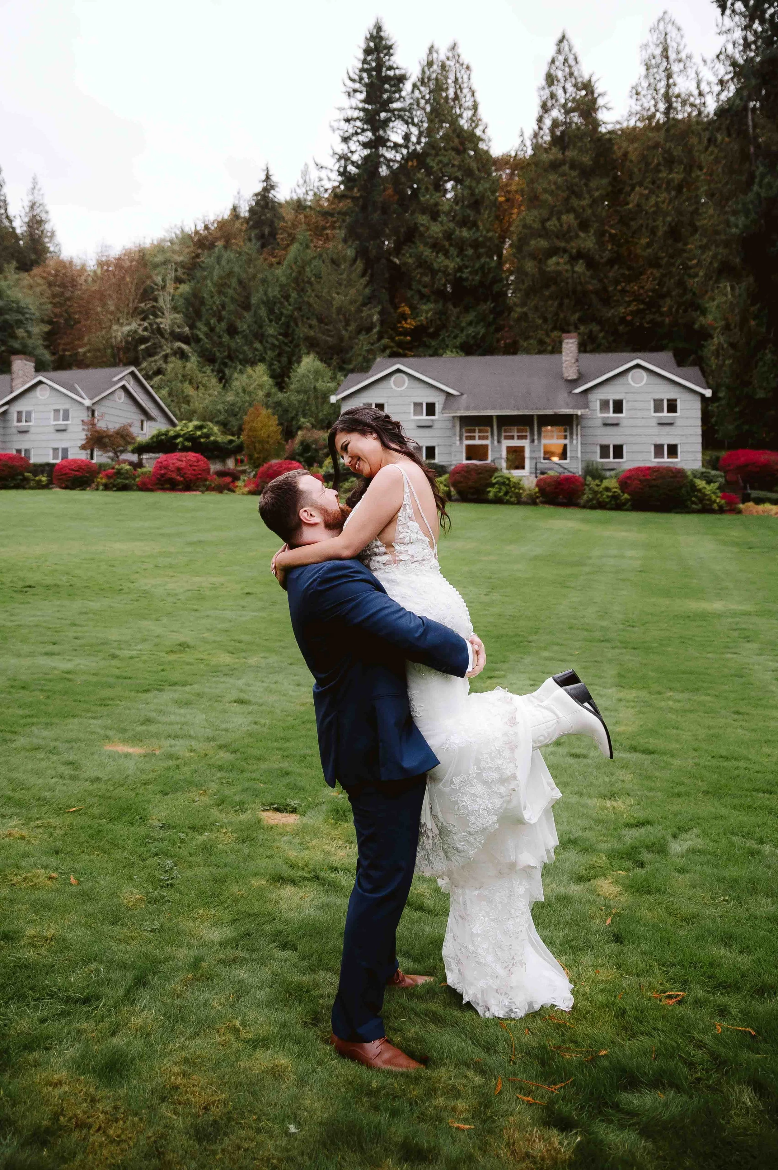 A happily married couple, with the man lifting the woman in white wedding dress, on lush green lawn with house and trees in the background.