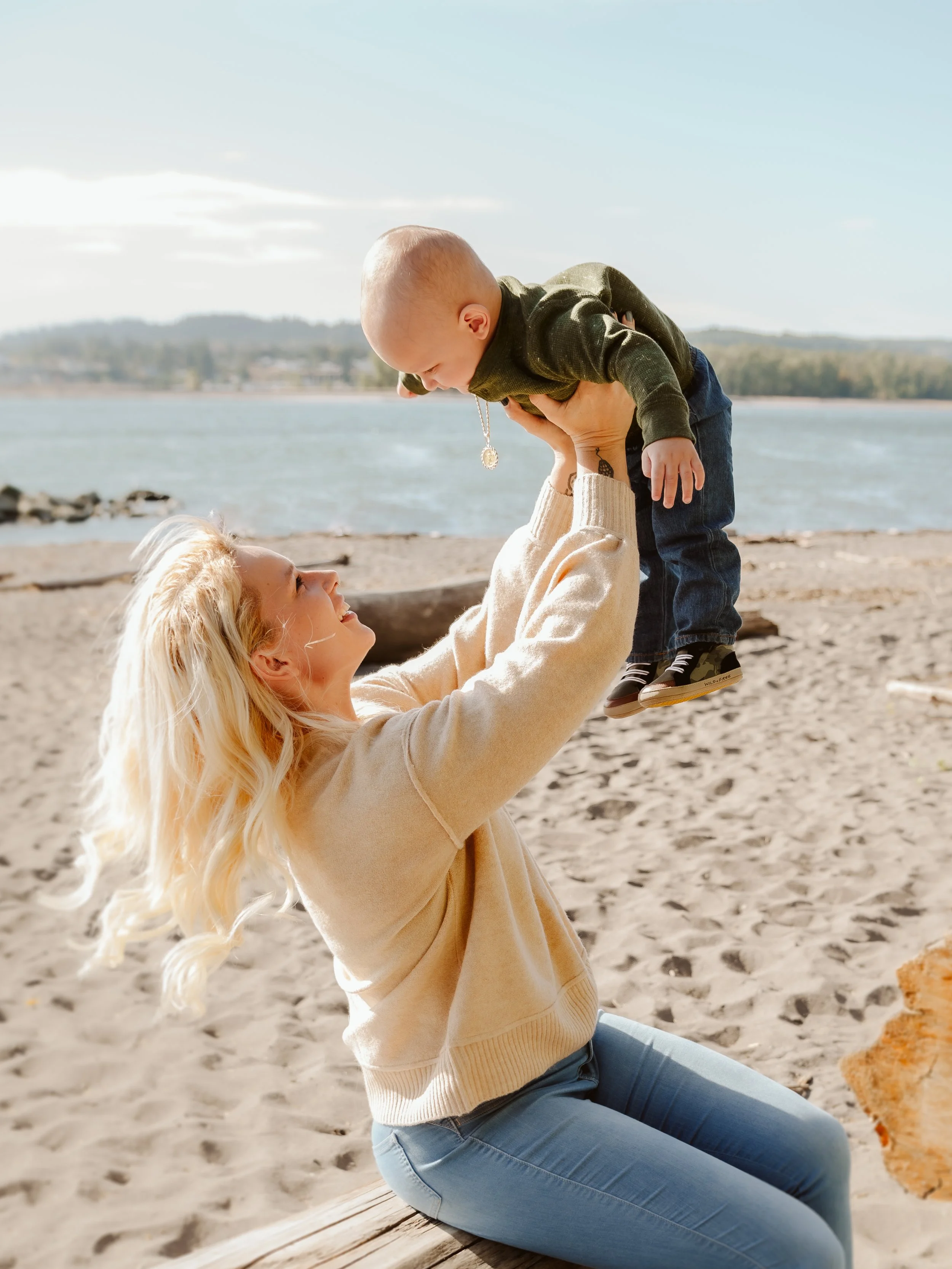 A woman with blonde hair playing with a young boy on a sandy beach near water, lifting him into the air and smiling at him.