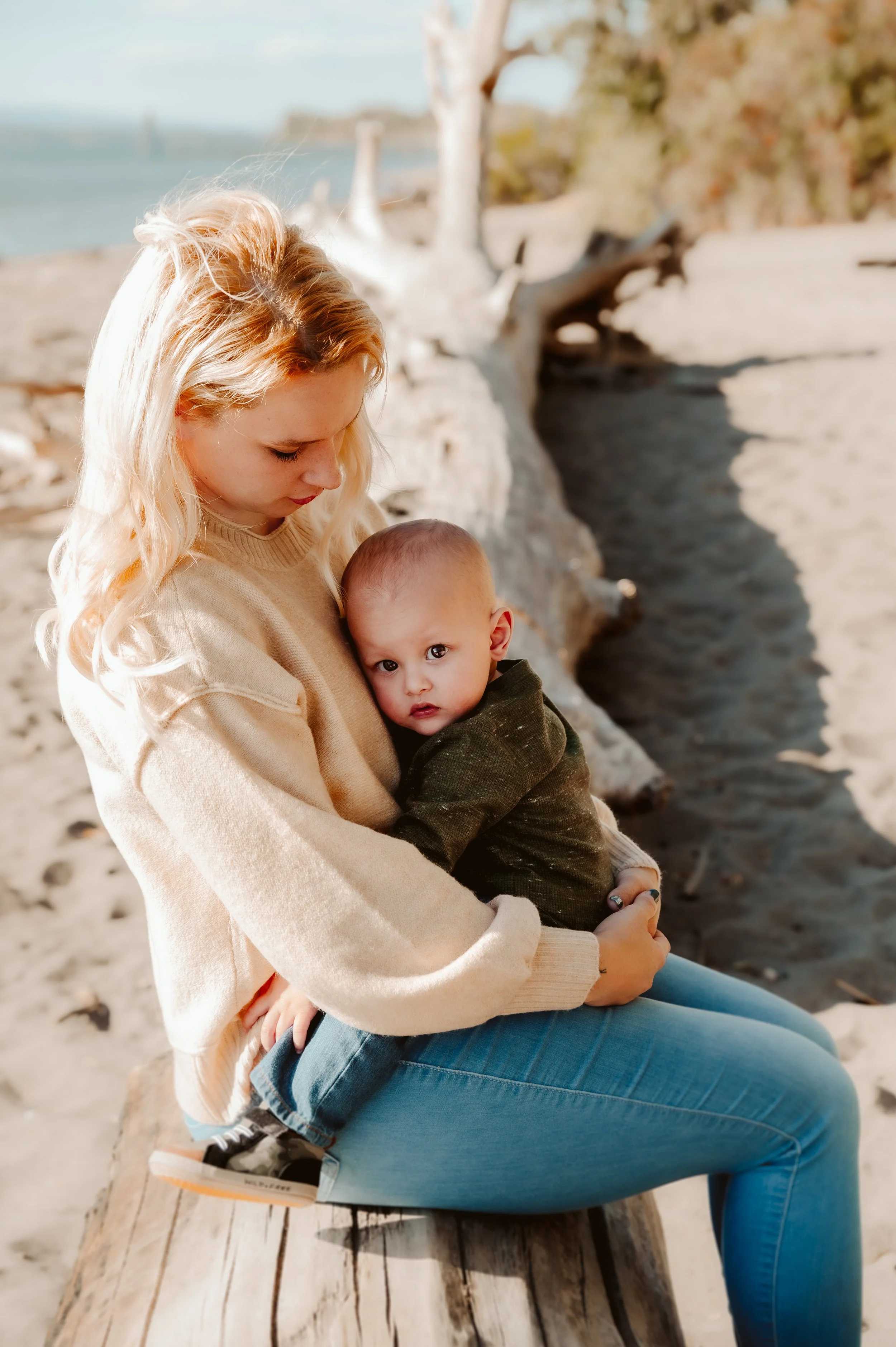 A woman with blond hair holding a young child with a shaved head on her lap on a sandy beach, with driftwood and trees in the background.