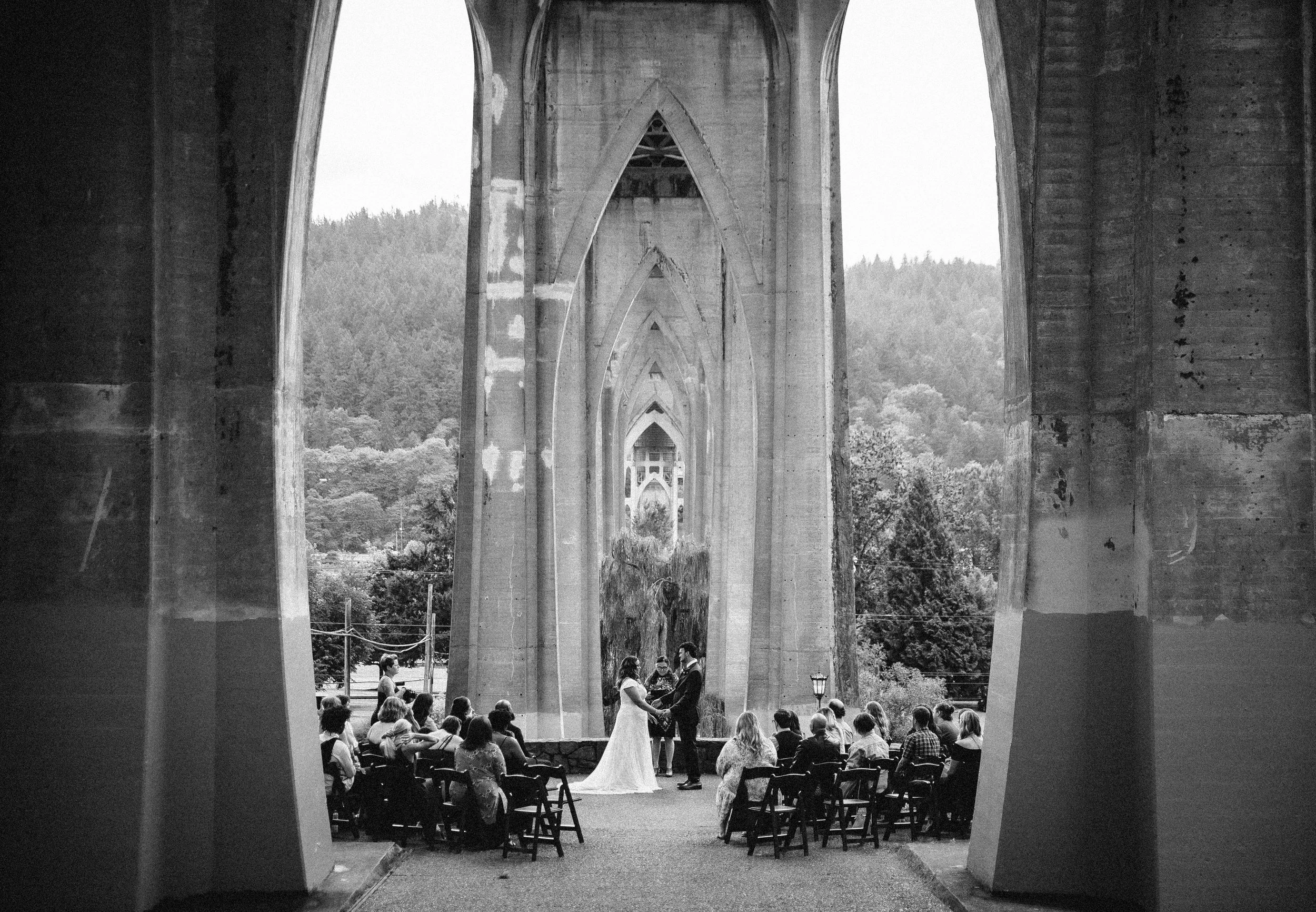 Bride and groom exchanging vows during a wedding ceremony beneath the St. Johns Bridge arches at Cathedral Park in Portland, Oregon.