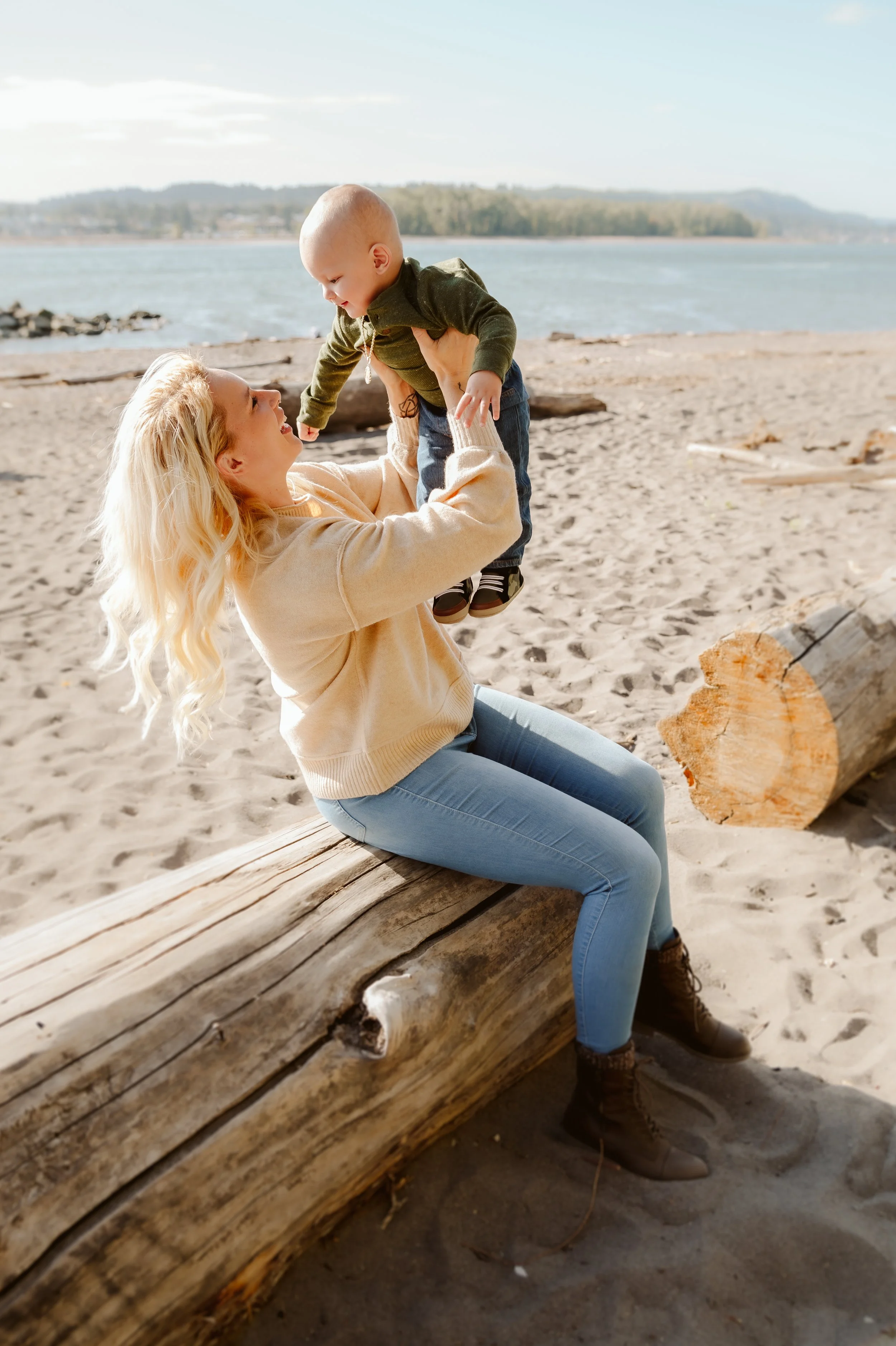 A woman with long blonde hair sitting on a large driftwood log on a sandy beach, lifting a young child with a shaved head and green sweater into the air, both smiling. The ocean and distant shoreline are visible in the background.