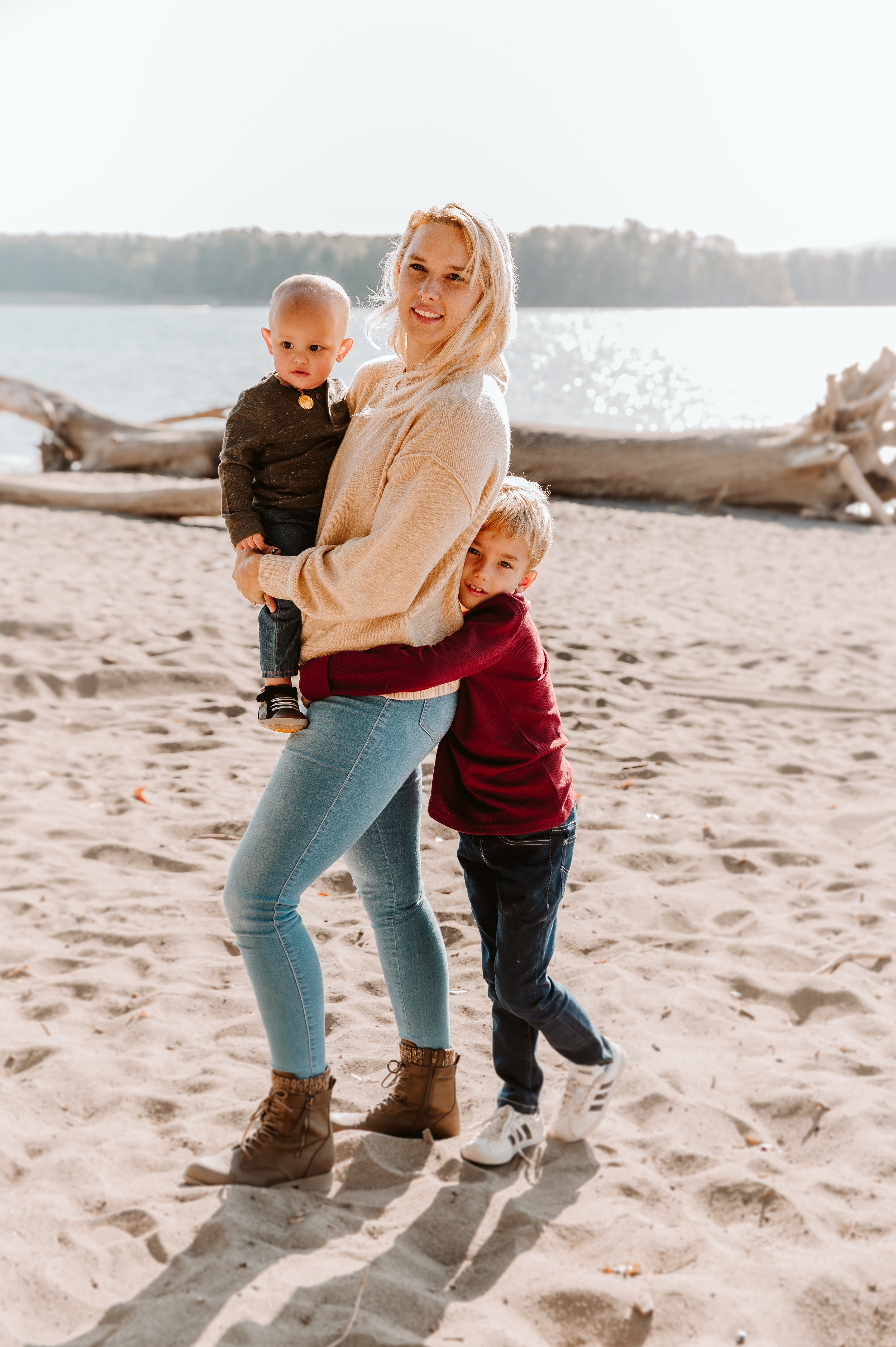 A woman with blonde hair standing on a sandy beach, holding a baby in her arms while a young boy clings to her waist. The background features driftwood and a body of water under a bright sky.