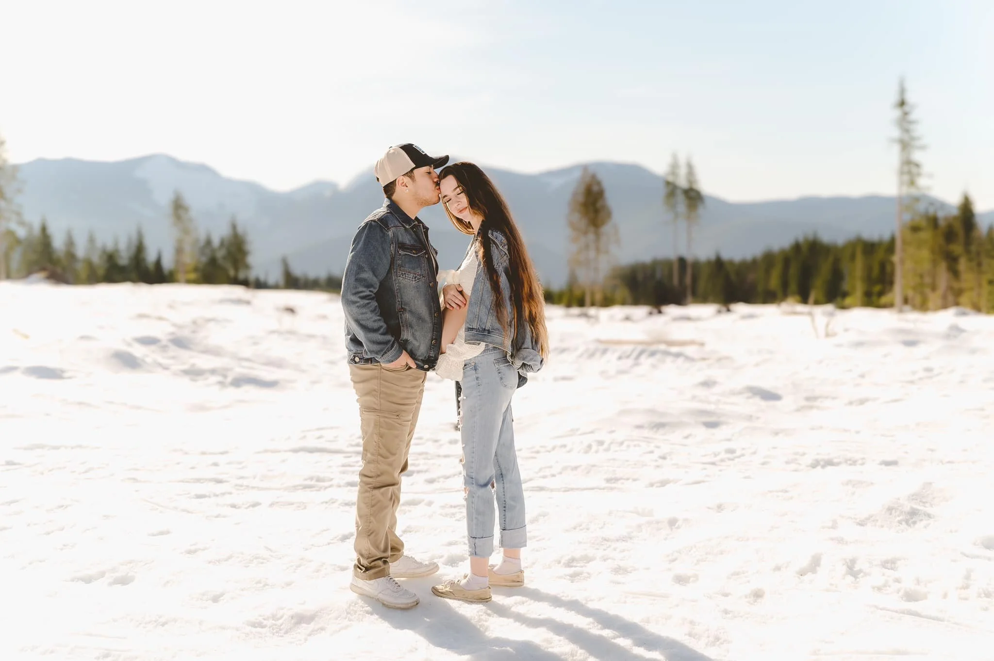 Young couple outdoors in a snowy landscape, with mountains and trees in the background, one kissing the other's forehead.