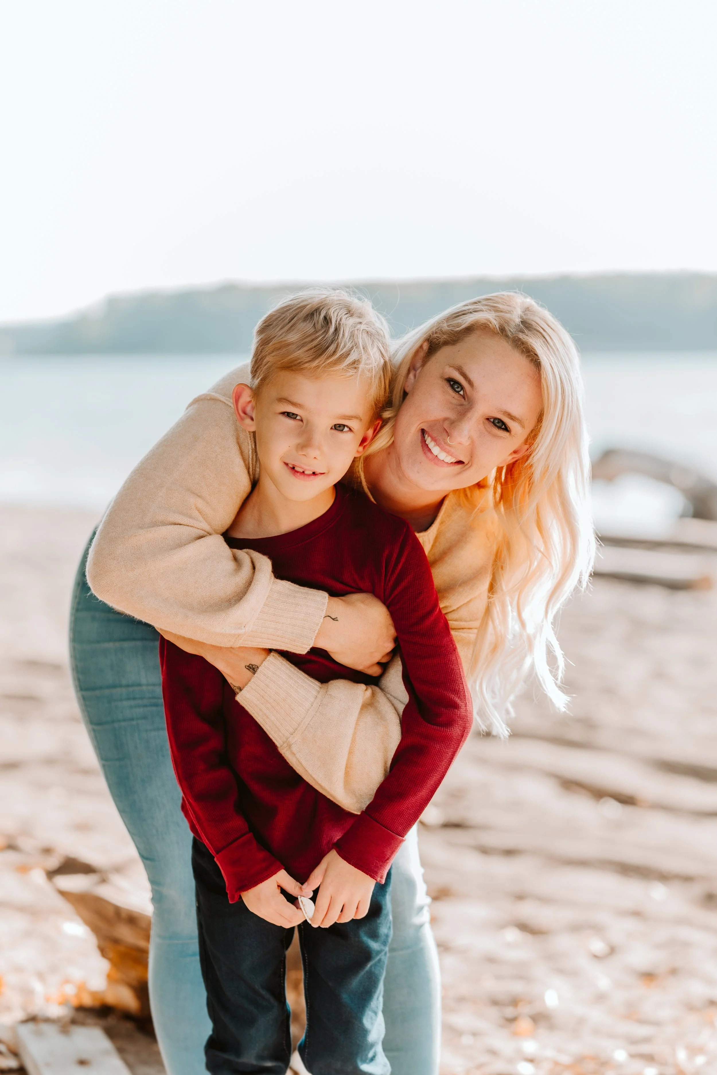 A woman and a young boy smiling and hugging at the beach with water and land in the background.
