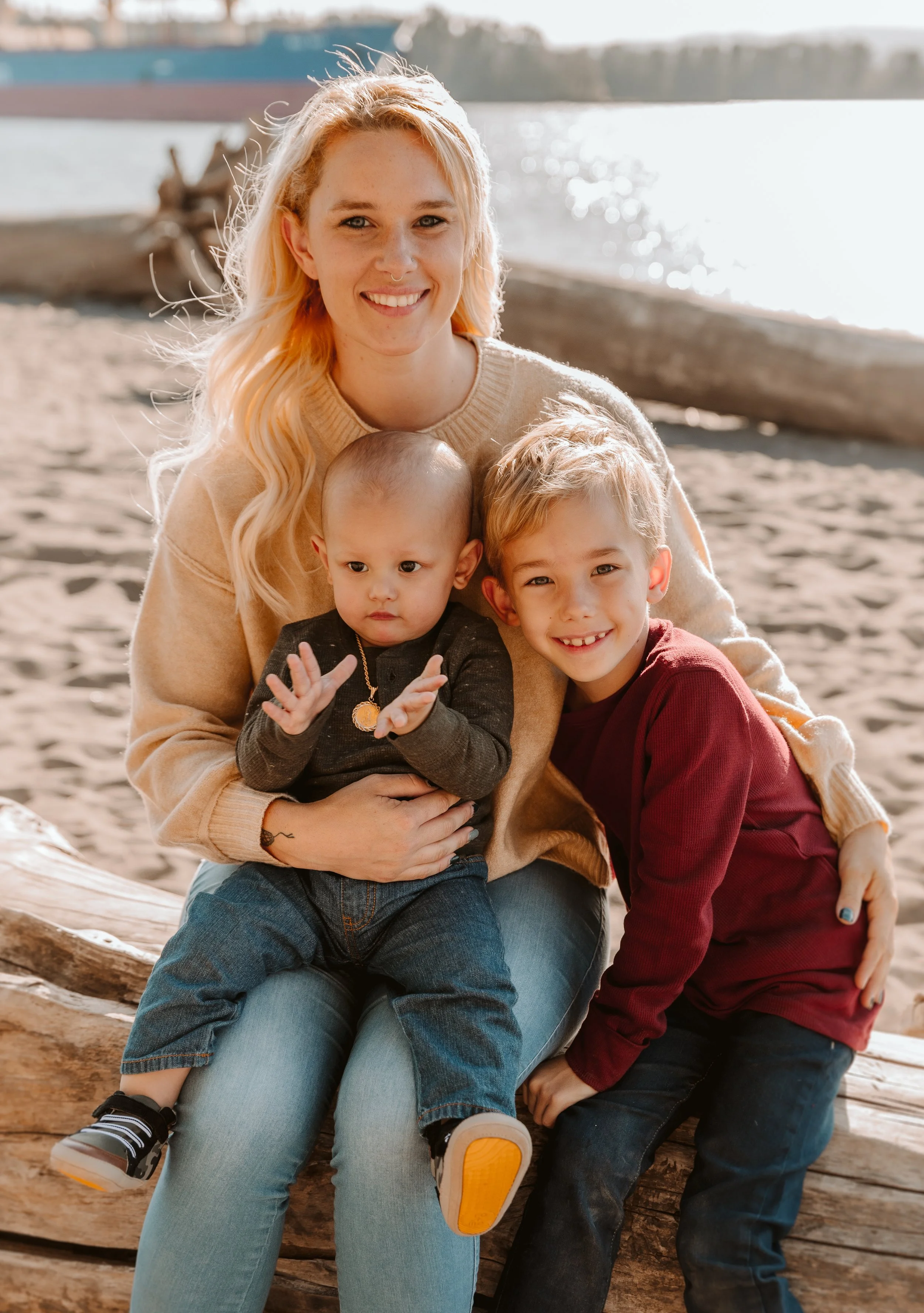A woman with long blonde hair and two young boys sitting on a large piece of driftwood at the beach, with water and a distant shoreline in the background.