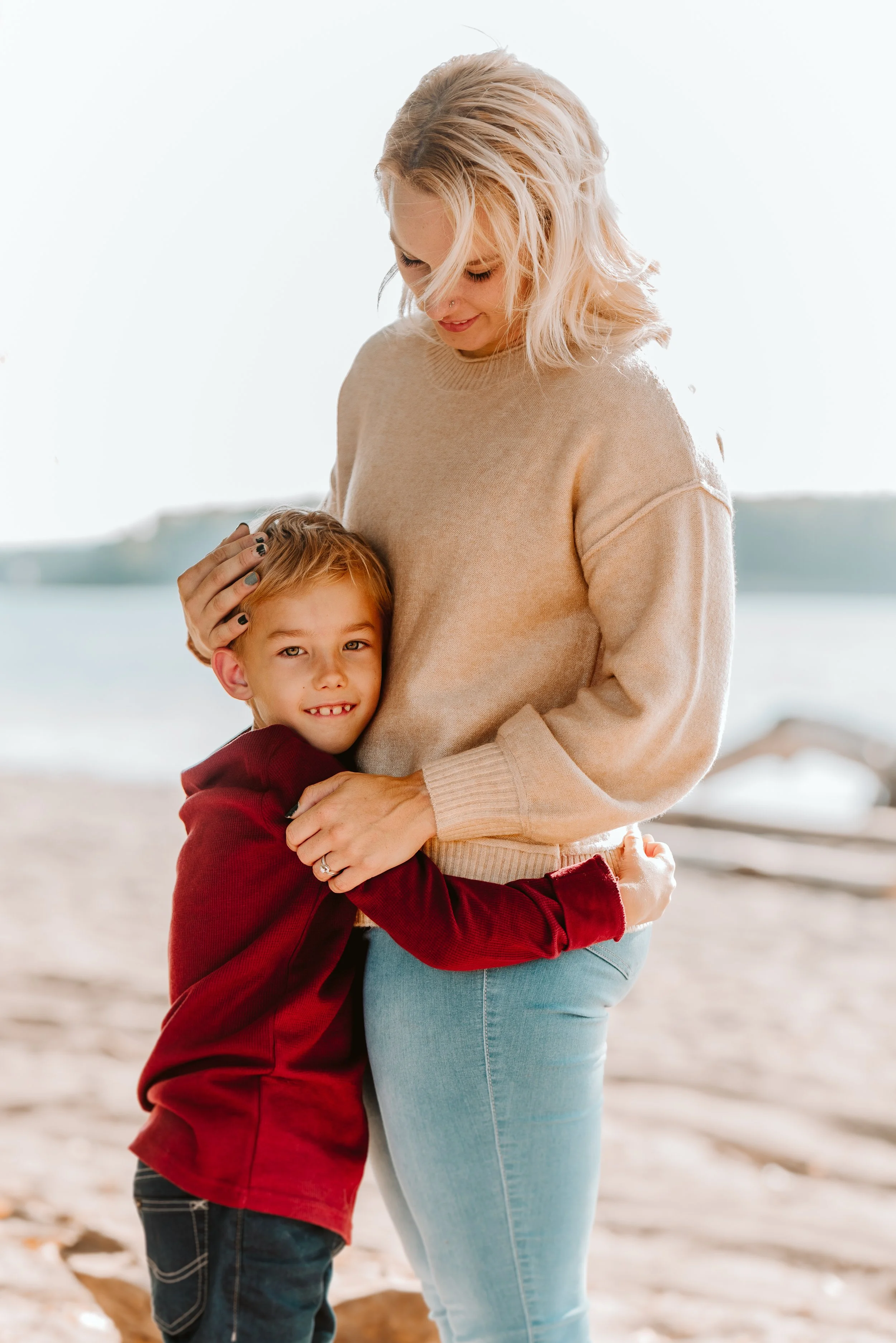 A woman and a young boy hugging on a beach near the water. The woman, with blonde hair, is wearing a beige sweater and light blue jeans. The boy, with light brown hair, is wearing a red hoodie and jeans, smiling at the camera.