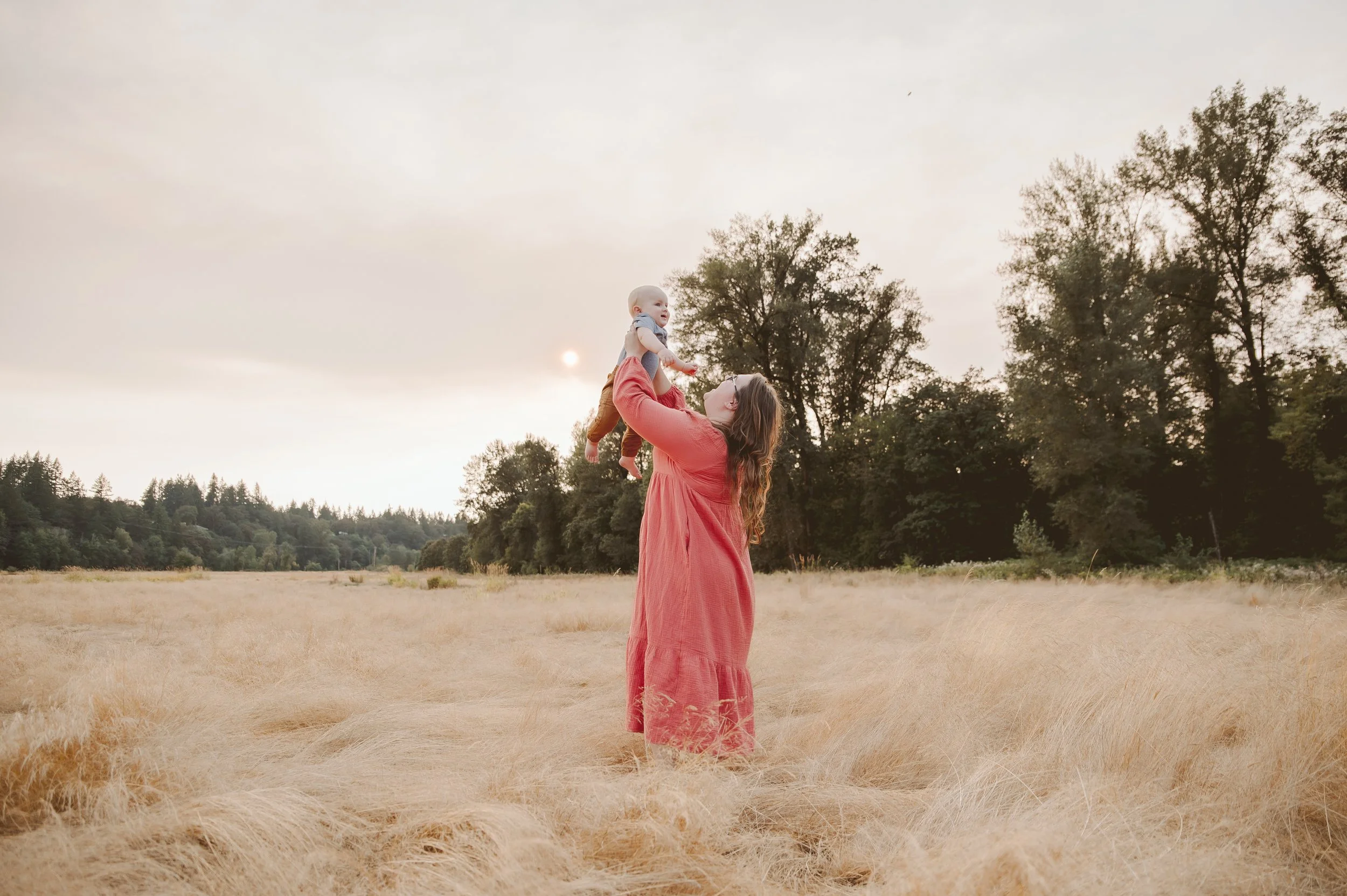 Golden hour motherhood photography of a woman lifting her baby in tall grass at sunset, captured by a Vancouver Washington family photographer.
