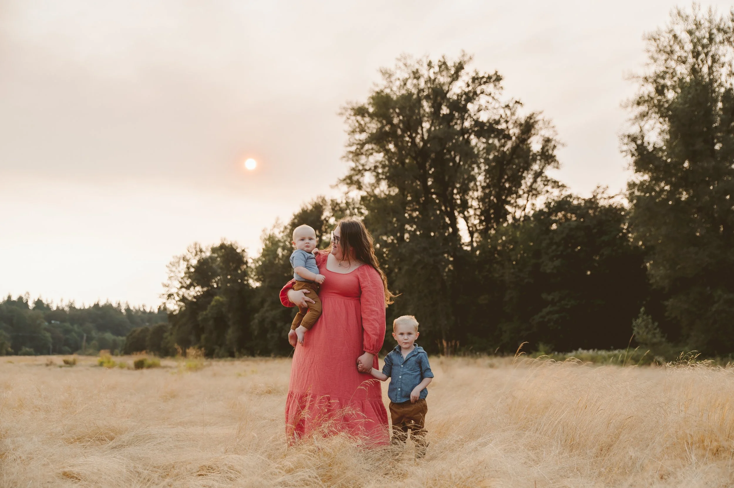 Motherhood and family photography of a woman holding two young children in tall golden grass during sunset, captured by a Vancouver Washington family photographer.
