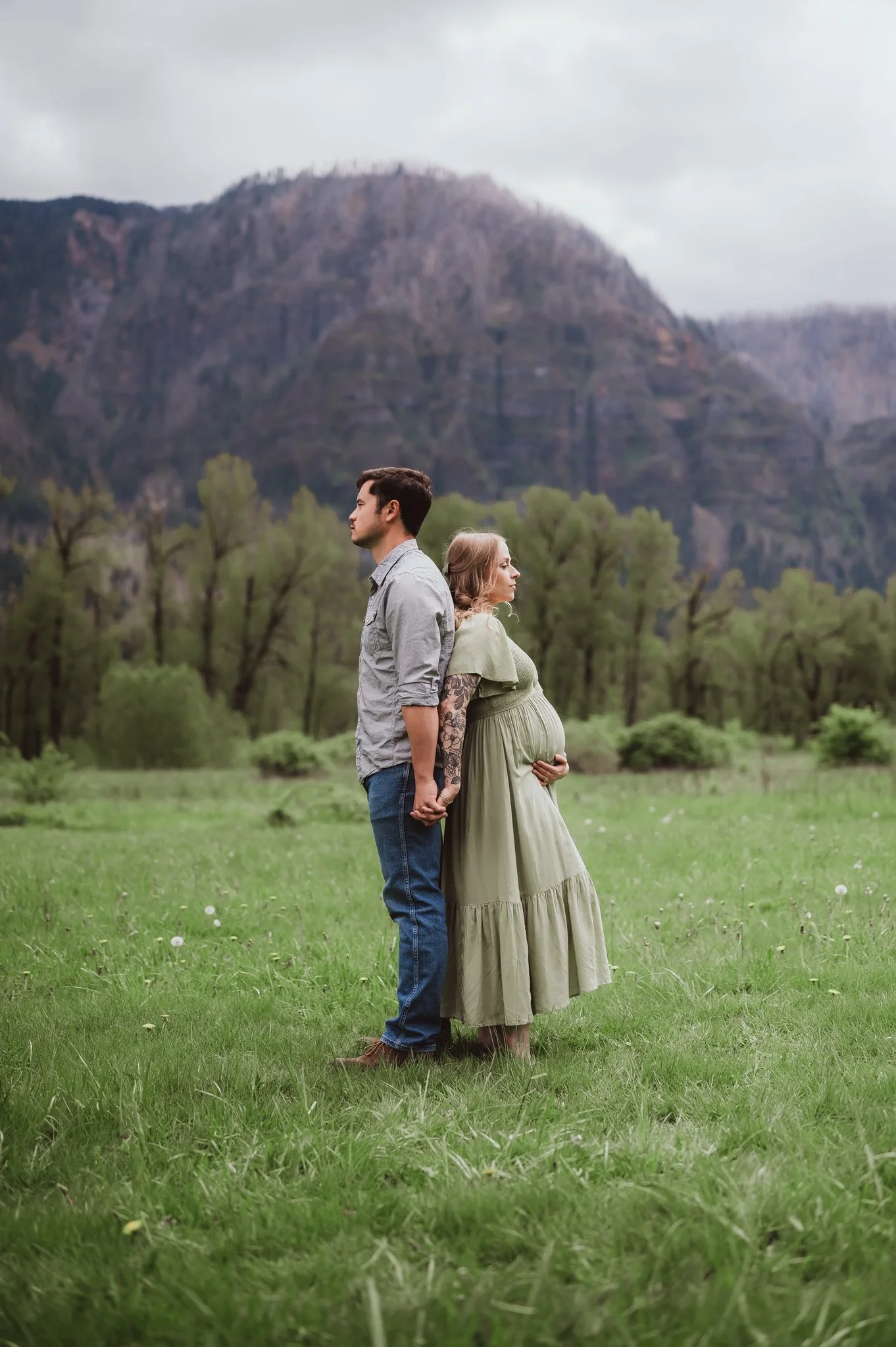 Outdoor maternity photo session in Vancouver, Washington featuring an expecting couple standing back to back in a grassy field with mountains and trees in the background, photographed by a local maternity photographer.