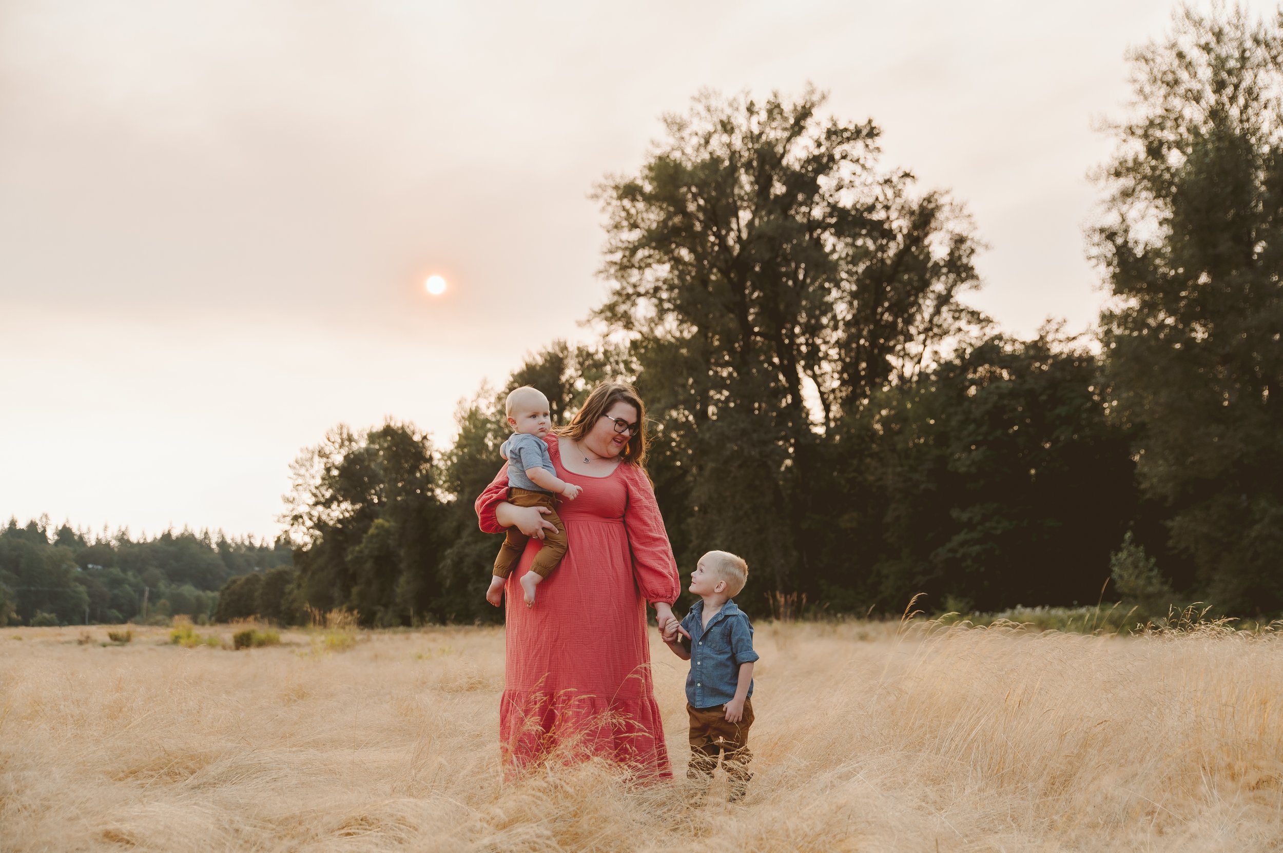 Lifestyle family portrait of a mother walking with her children through a sunlit field of tall grass at sunset, photographed by a Vancouver Washington photographer.
