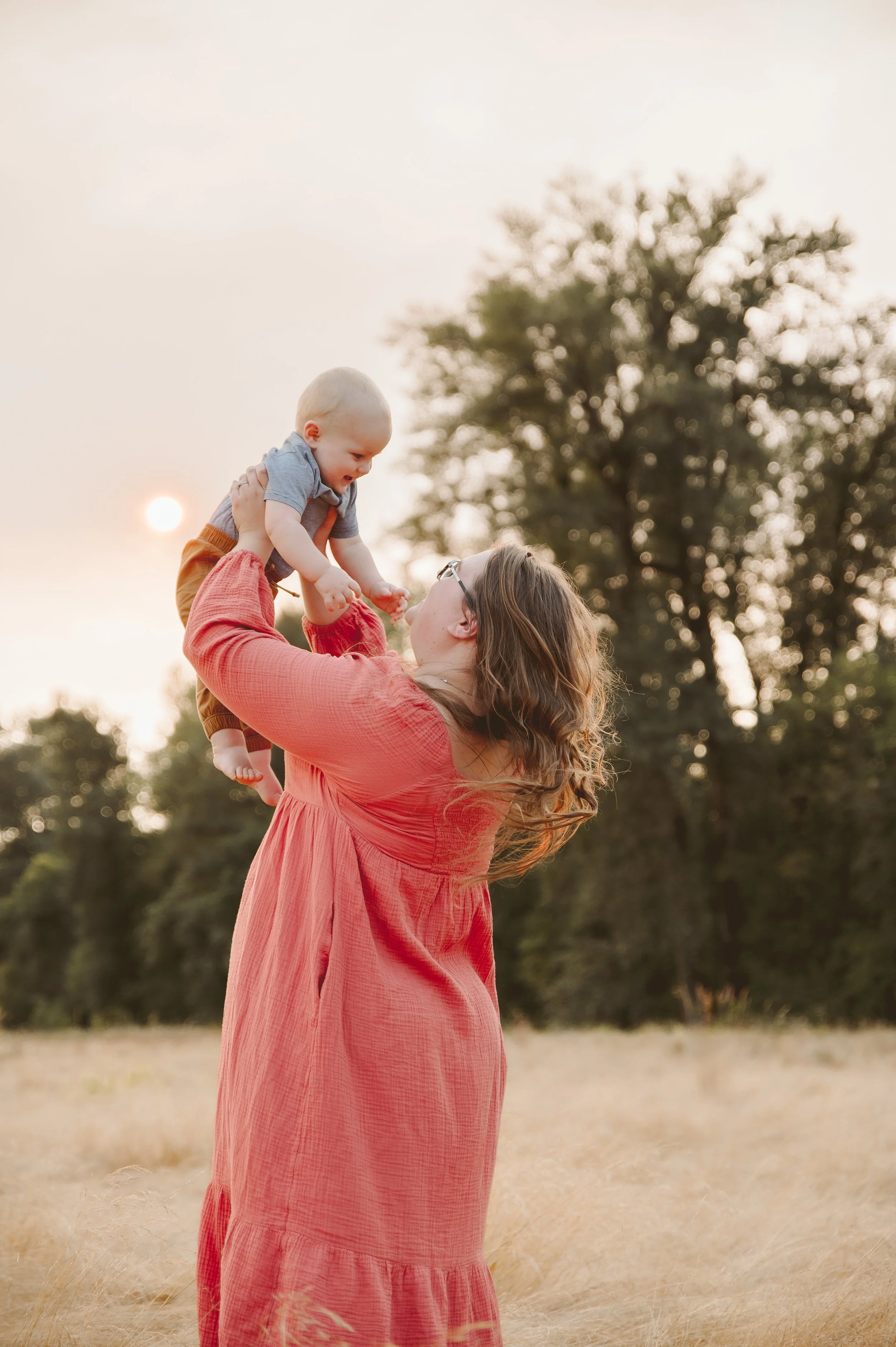 Lifestyle motherhood photography of a woman lifting her baby during golden hour in an open field, photographed by a Vancouver Washington photographer.
