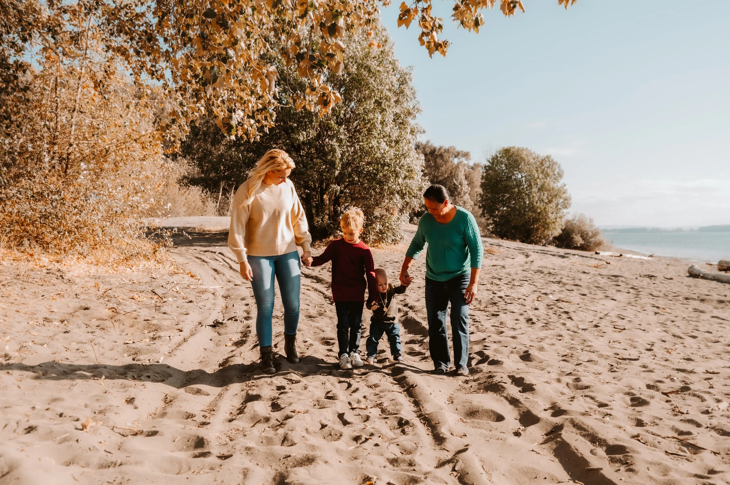 Two women and two children walk along a sandy beach holding hands on a sunny autumn day, with trees and water in the background.
