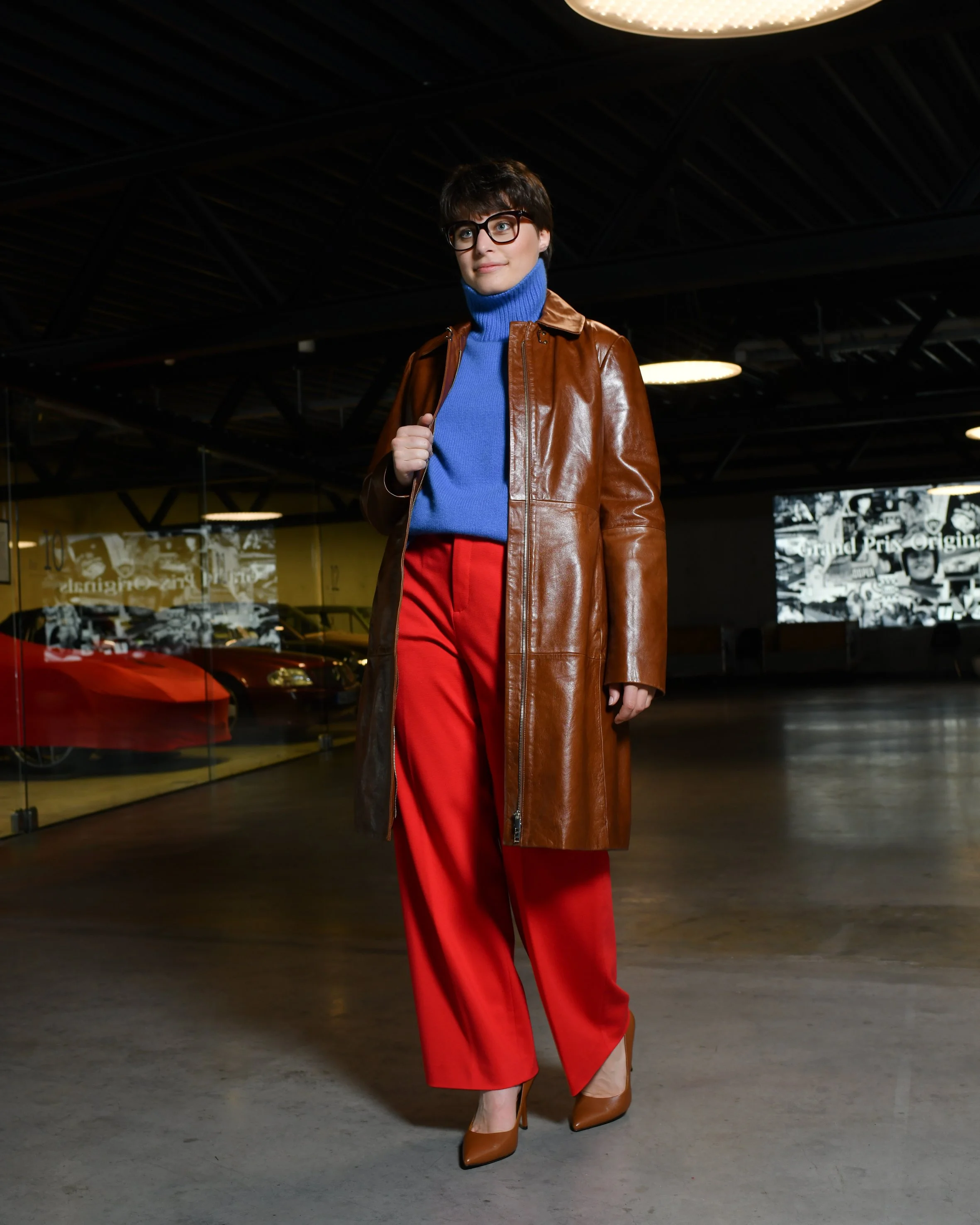 Frau mit kurzen braunen Haaren. Sie trägt eine lange rote Hose und einen blauen Rollkragenpullover. Darüber trägt sie einen langen braunen Ledermantel und braune Keilabsatzschuhe.