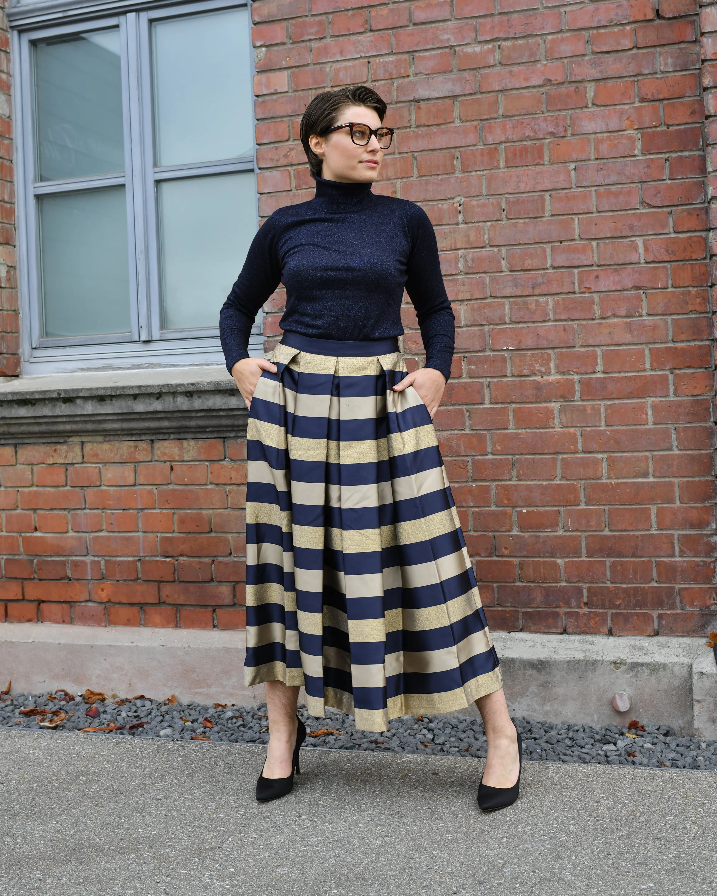 Eine Frau mit trägt einen langen gestreiften Rock in gold, dunkelblau. Dazu ein dunkelblaues Langarmshirt und schwarze elegante Absatzschuhe.