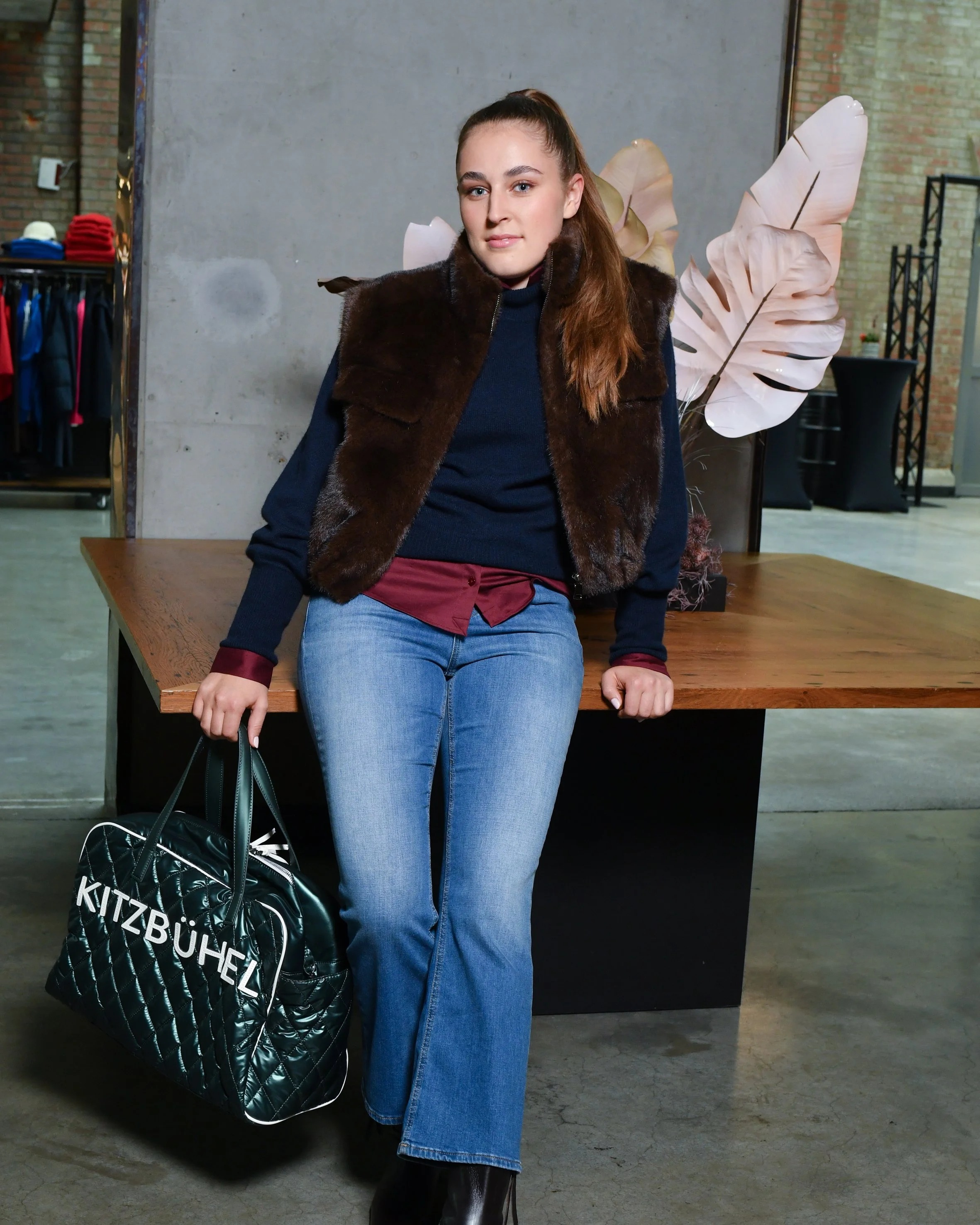 Frau mit braunen langen Haaren. Sie trägt eine hellblaue Jeans, eine dunkelrote Bluse und darüber einen dunkelblauen Pullover sowie eine kurzärmliche braune Weste. Sie hält eine türkis-grüne Tasche in der Hand, auf der Kitzbühel steht.