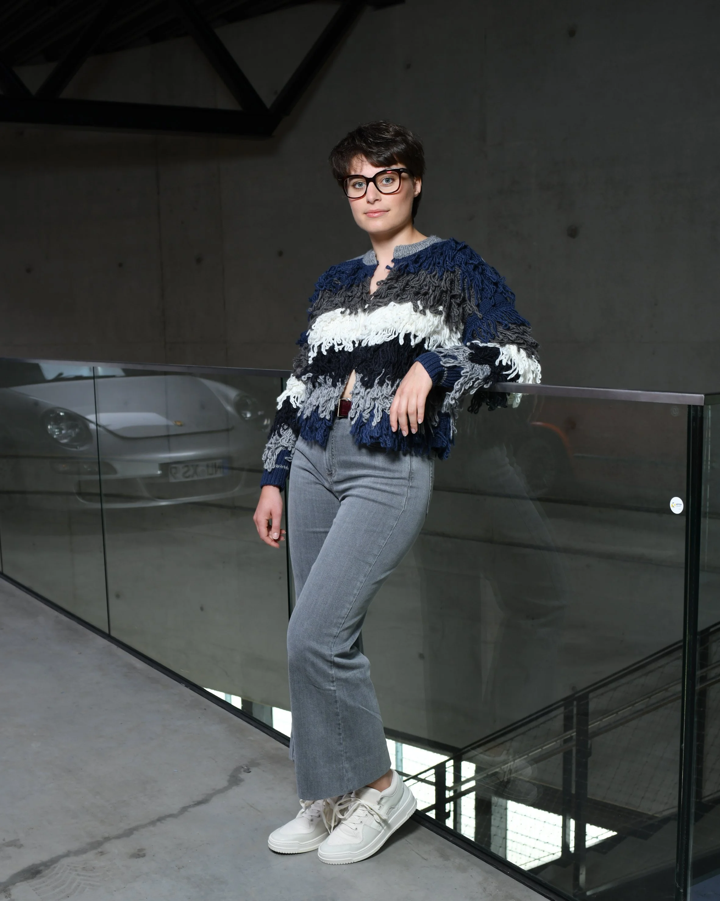 Frau mit kurzen braunen Haaren. Lehnt an einem Glasgeländer. Sie trägt eine graue Jeans und ein Gestreiftes Franzenoberteil in den Farben: olive, weiß, dunkelblau, grau. Dazu trägt sie weiße Sneaker.