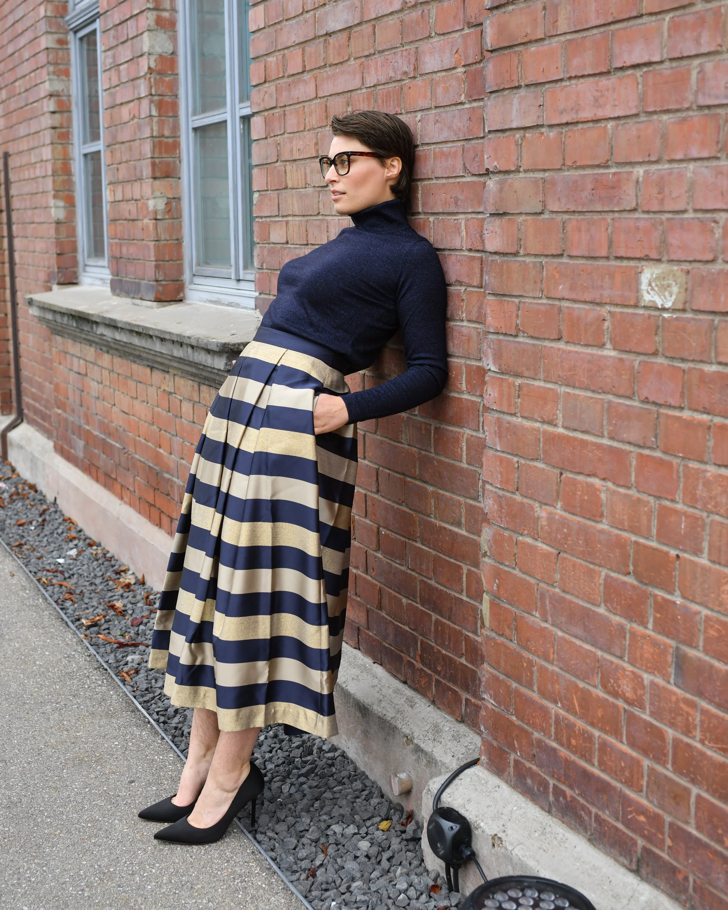 Eine Frau mit trägt einen langen gestreiften Rock in gold, dunkelblau. Dazu ein dunkelblaues Langarmshirt und schwarze elegante Absatzschuhe.