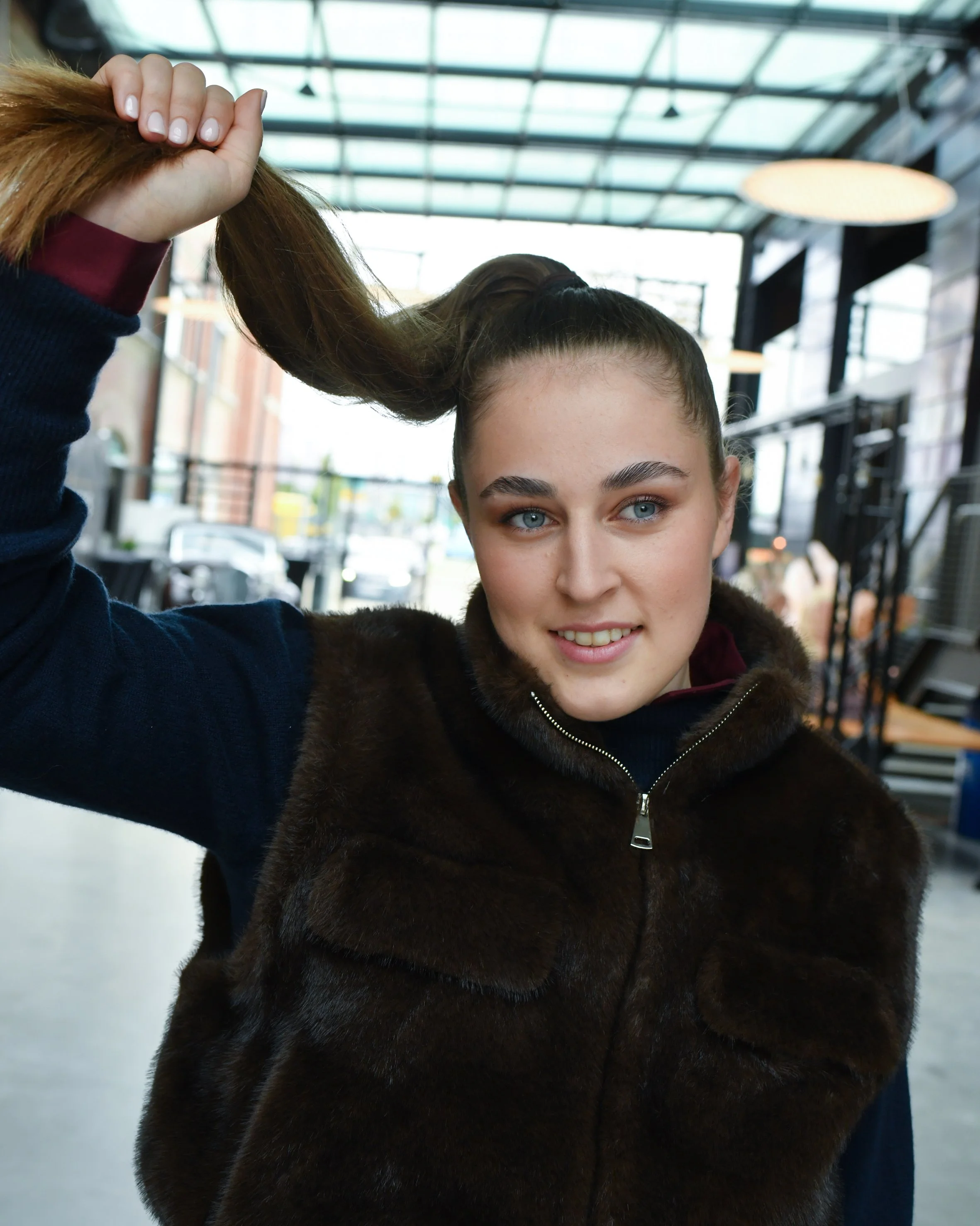 Frau mit braunen langen Haaren. Sie trägt eine dunkelrote Bluse und darüber einen dunkelblauen Pullover sowie eine kurzärmliche braune Weste. 