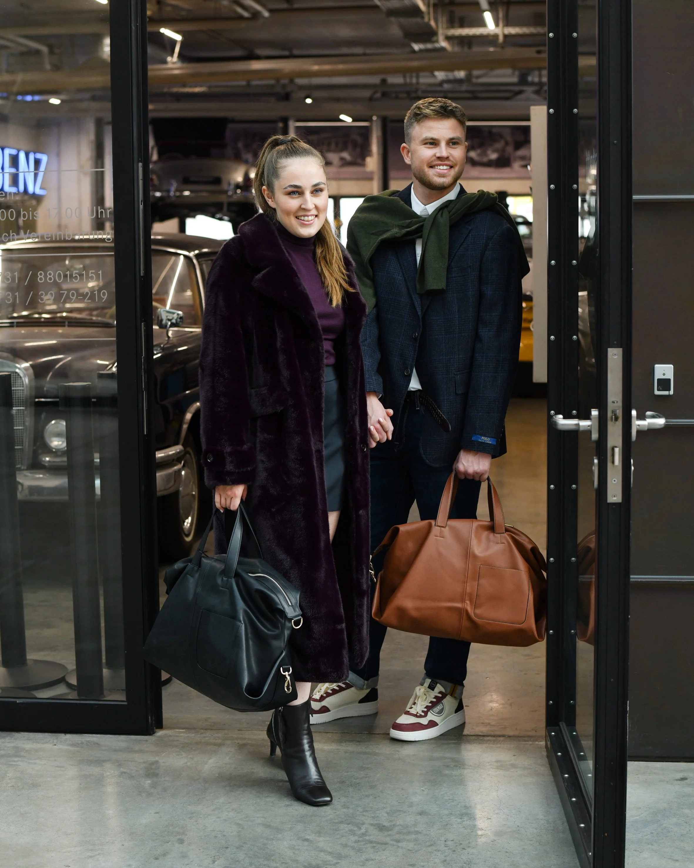 Eine Frau und ein Mann, händchen haltend. Die Frau trägt einen dunkelblauen kruzen Rock und ein bordeau rotes Rollkragensweathshirt, dazu einen langen kuscheligen bordoufarbenen Mantel. Zudem trägt sie einen große schwarze Tasche und Absatz Stifelett
