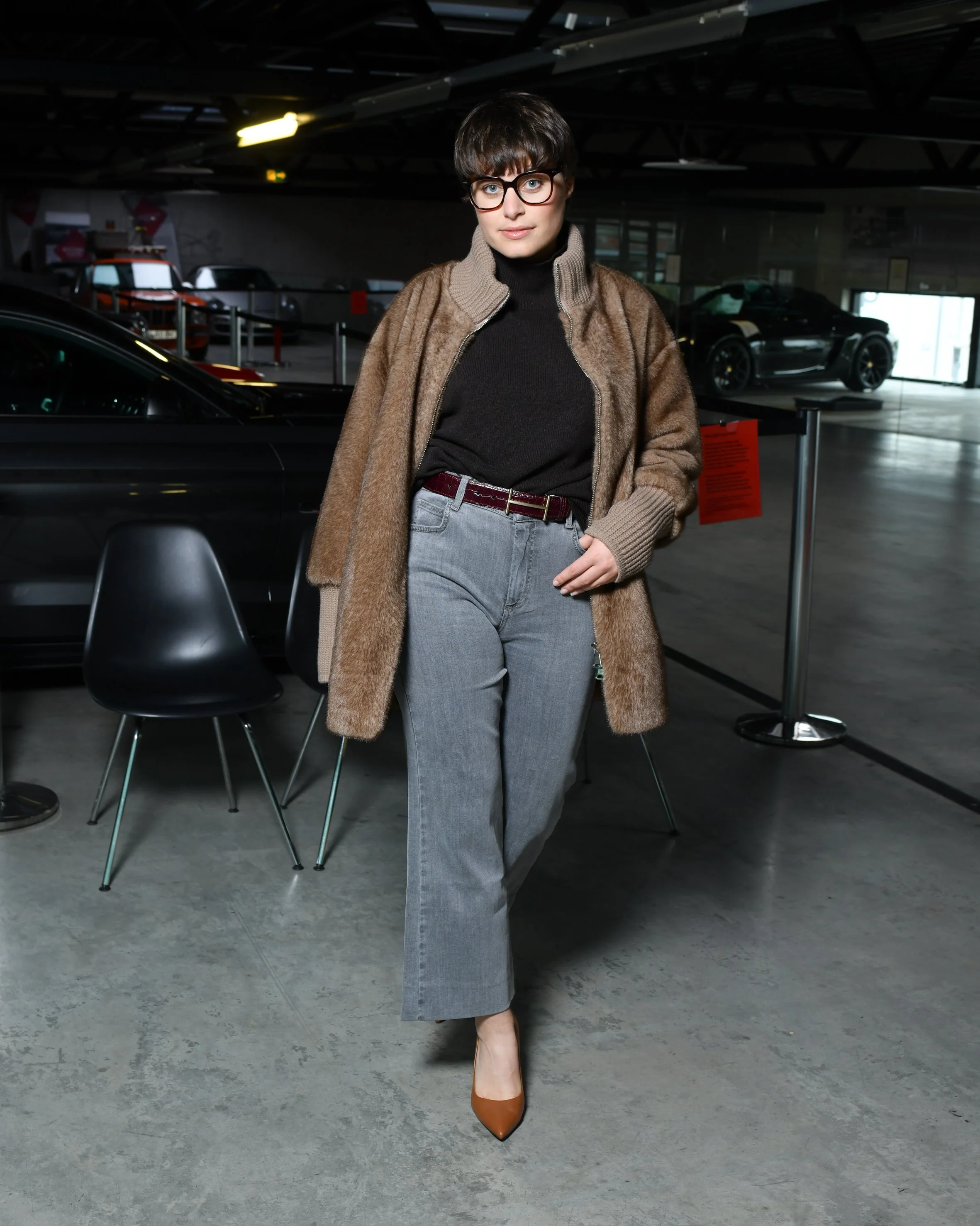 Frau mit grauer, weiter Jeans. Schwarzer Pullover und darüber eine braune kuschelige Jacke. Hohe Schuhe in braun. Sie steht und lächelt in die Kamera.