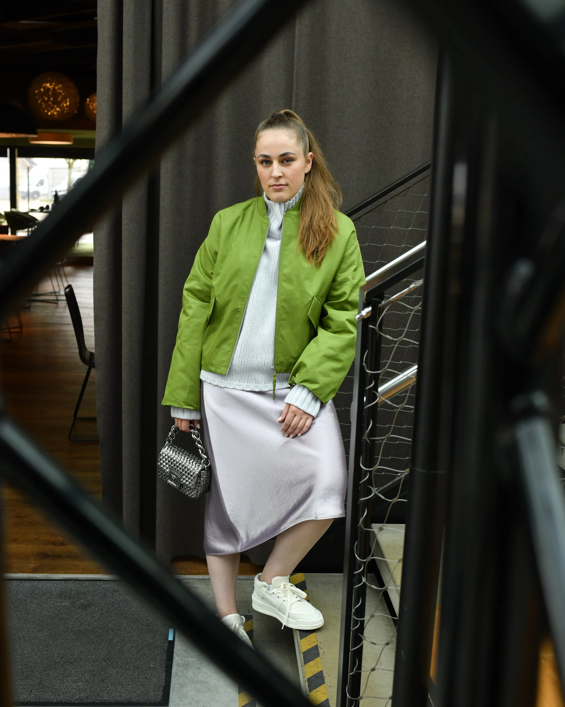 Frau mit braunen langen Haaren. Sie trägt einen Seidenrcok in rose, dazu einen grauen Rollkragenpullover und eine grüne Bomberjacke. Dazu trägt sie eine silberne kleine Handtasche und weiße Sneaker