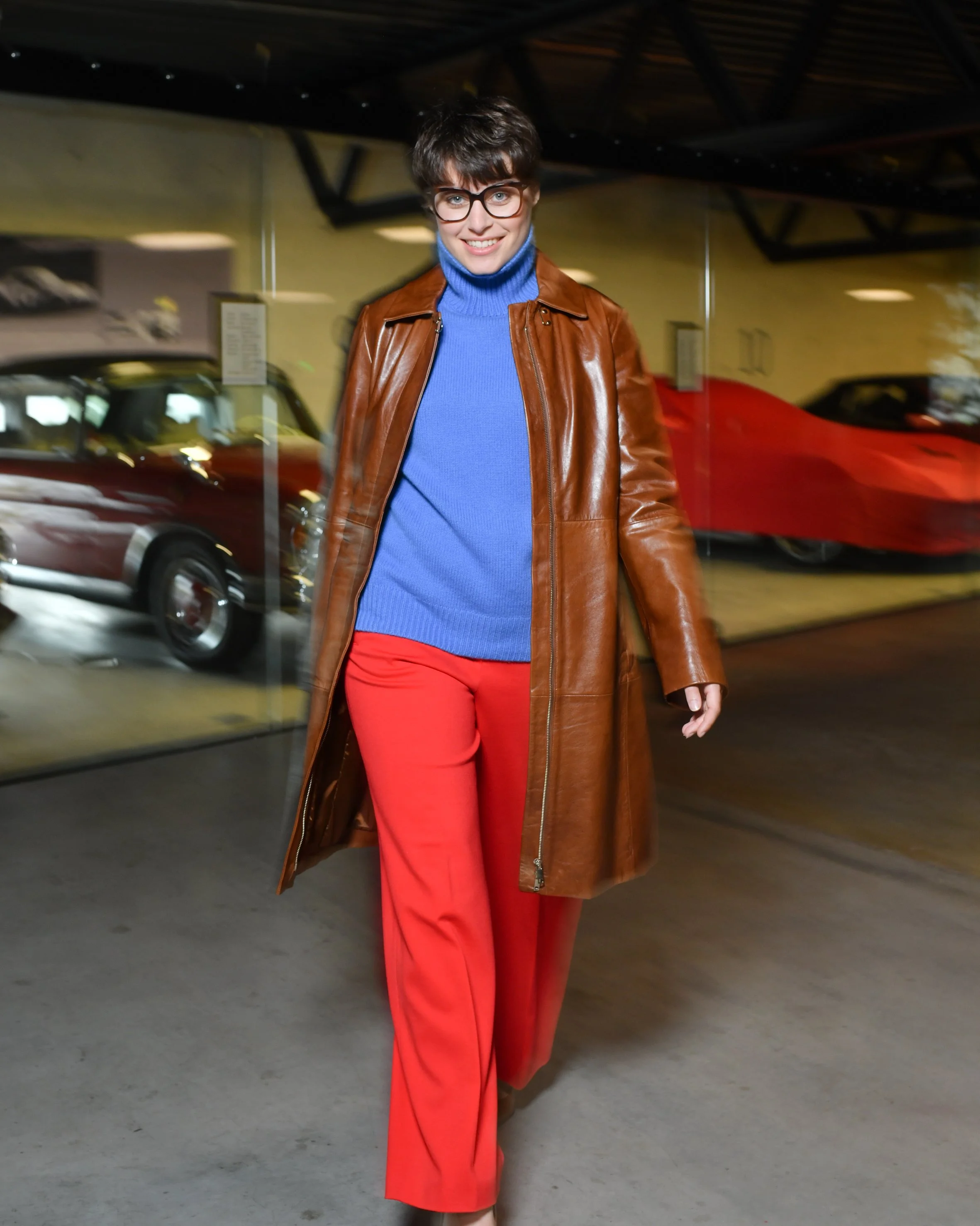 Frau mit kurzen braunen Haaren. Sie trägt eine lange rote Hose und einen blauen Rollkragenpullover. Darüber trägt sie einen langen braunen Ledermantel und braune Keilabsatzschuhe.
