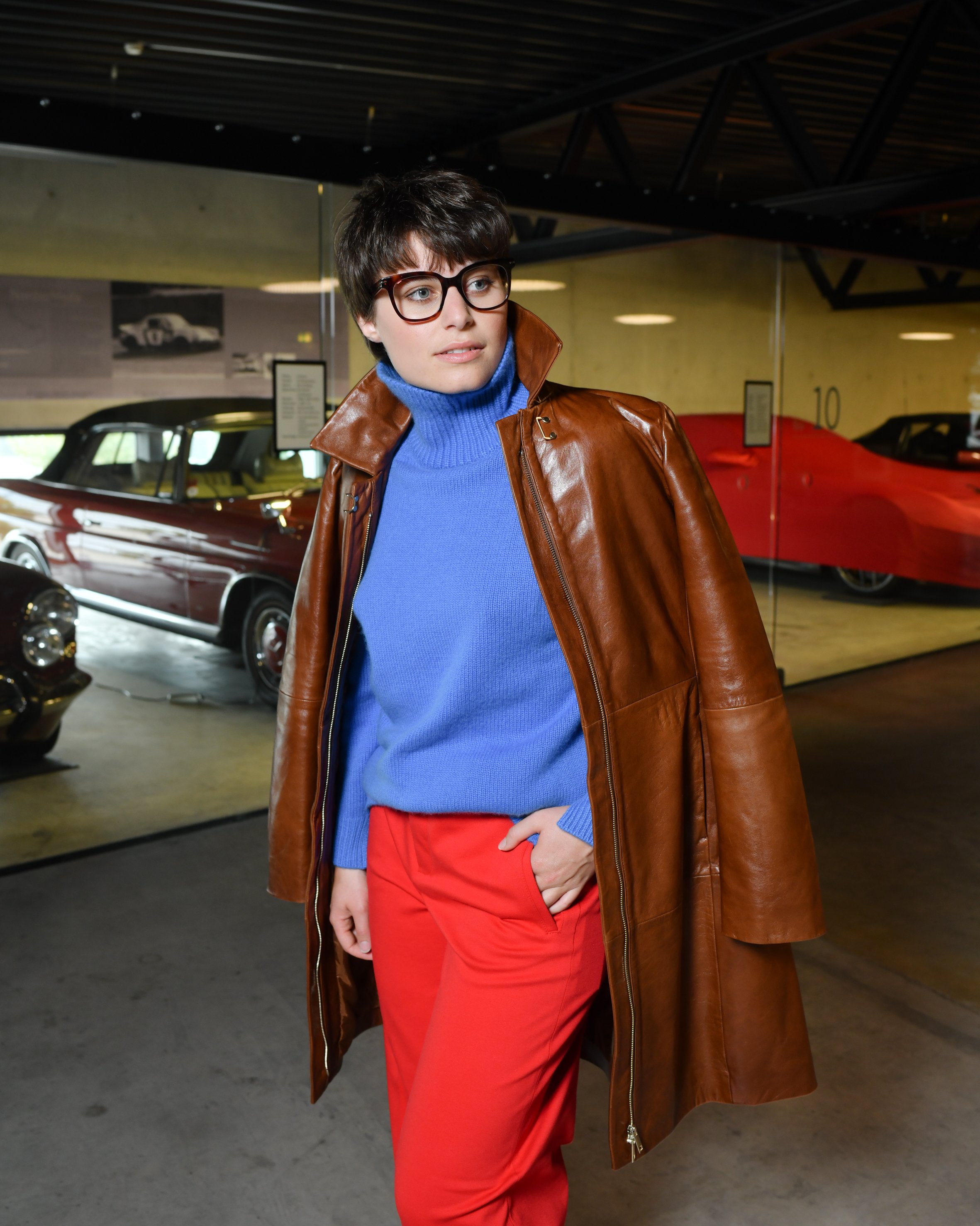 Frau mit kurzen braunen Haaren. Sie trägt eine lange rote Hose und einen blauen Rollkragenpullover. Darüber trägt sie einen langen braunen Ledermantel und braune Keilabsatzschuhe.